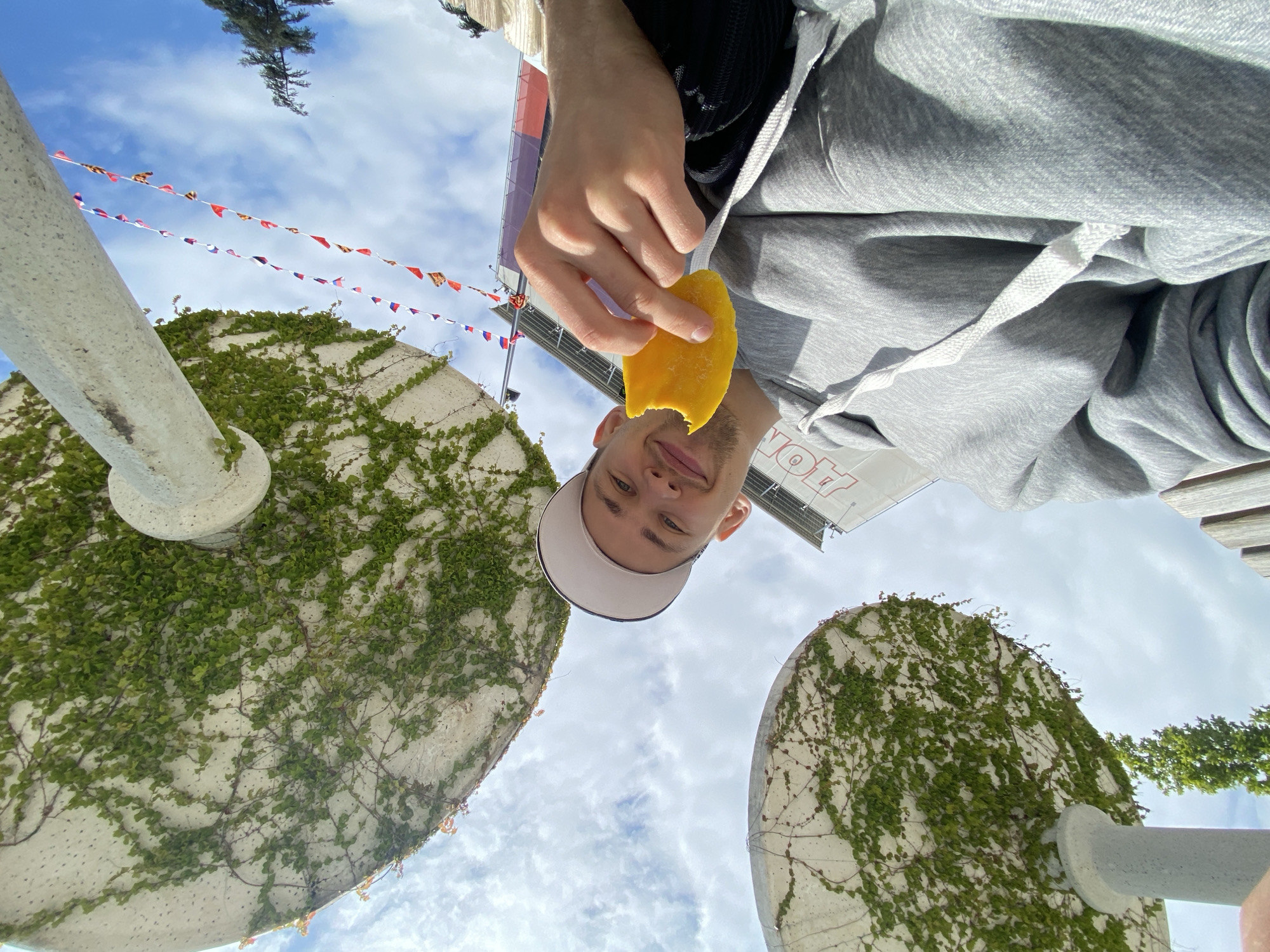 The image depicts a person holding a piece of mango and looking towards the camera, taken from a low angle pointing upwards. The person wears a white baseball cap and a grey hoodie. A bite has been taken out of the mango. In the background are two circular, concrete structures covered in green ivy, positioned on either side of the person. Above the structures, multicolored pennant flags hang, strung between poles. The sky is visible through the gaps, revealing a mix of blue sky and white clouds. The location is город Ялта, Ukraine.