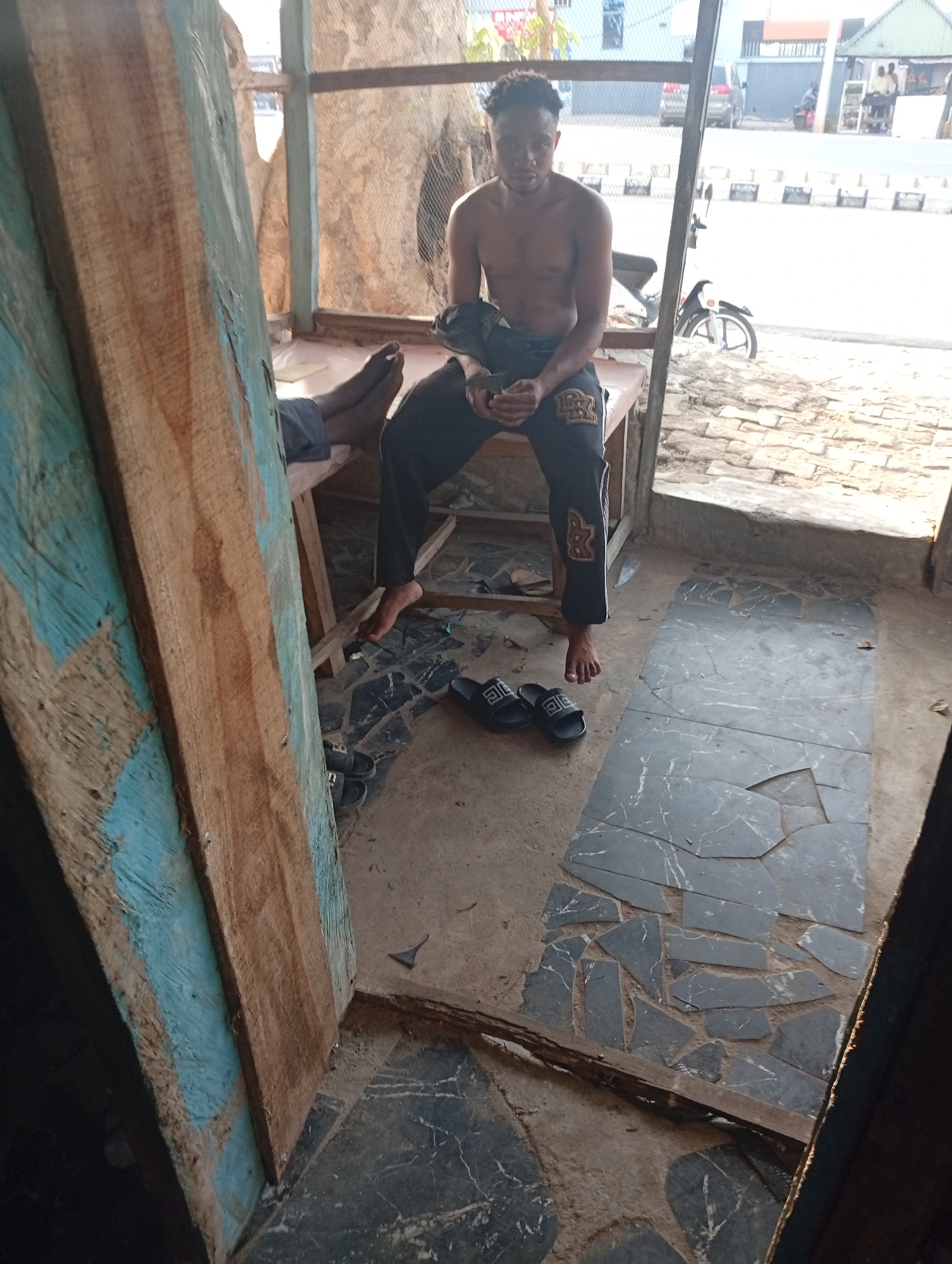 A shirtless man sits on a bench inside a rough, open-air structure. He is dark-skinned and wears dark pants. Another person is sitting on the same bench, with their foot visible. The ground is a mix of dirt and broken tiles. Outside the structure is a street scene with a parked motorcycle and other buildings. The image was taken in Adankolo, Nigeria.
