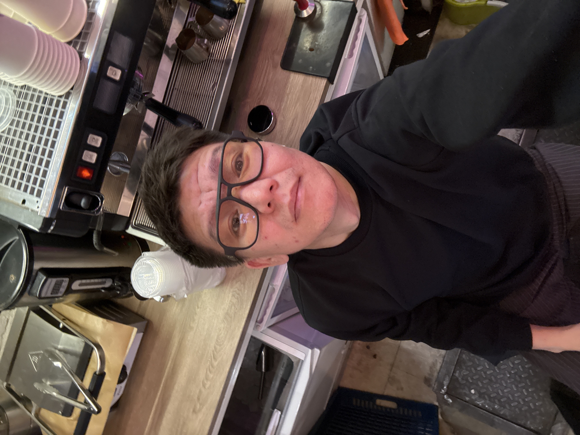 A person is taking a selfie in a cafe. They are wearing a black sweatshirt and glasses. The background shows an espresso machine, stacked cups, and a metal grate on the floor. The person's location is not specified, but the context suggests a commercial establishment.