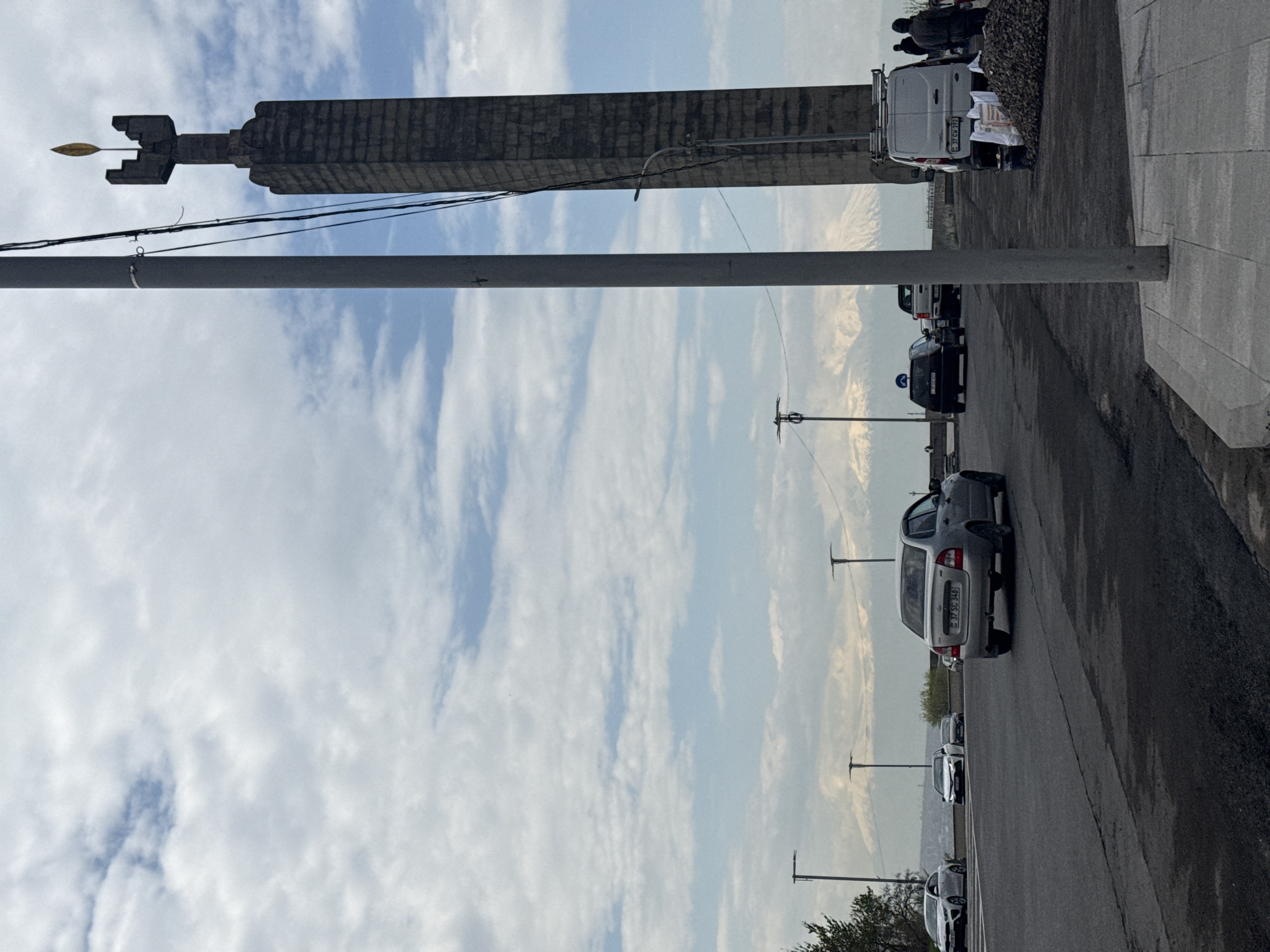 The image shows the Yerevan Genocide Memorial in Armenia. A tall, gray stone structure, likely part of the memorial, dominates the top of the frame. A metal pole runs vertically in the foreground. The sky is partly cloudy with white streaks. Below the memorial, there's a street with parked cars. Some people are near a van, which is likely related to the memorial site. The street has a sidewalk on the right, and a mountains in the distance, hazy due to atmospheric perspective. The photo is taken from a slightly low angle, looking up at the memorial.