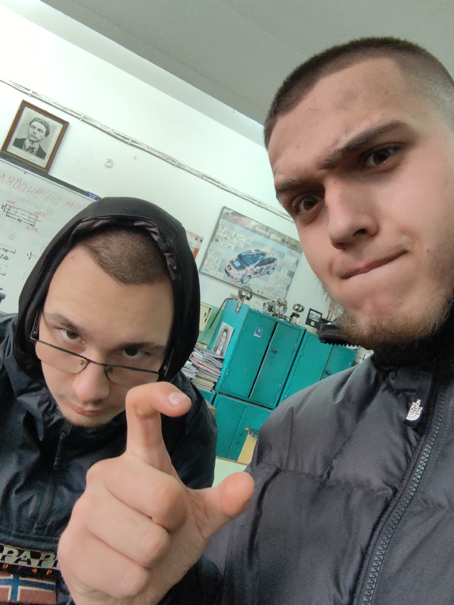 Two young men are in what appears to be a classroom or office setting. The man on the left wears a black hooded jacket and glasses and has a quizzical expression. He is making a small pinching gesture with his hand. The man on the right wears a black jacket and has a close-cropped haircut. He has a slight frown and seems to be looking directly at the camera.
Behind them, there is a whiteboard with writing on it, a framed portrait of a man, and a poster of a car on the wall. In the background, there are green filing cabinets. The jackets worn by both the men have branding labels on them which may help identify the location or time this image was taken.
While it is not possible to confirm the location in Sofia, Bulgaria, without further information, the presence of Cyrillic script on the whiteboard and the framed portrait could indicate a location in Bulgaria.