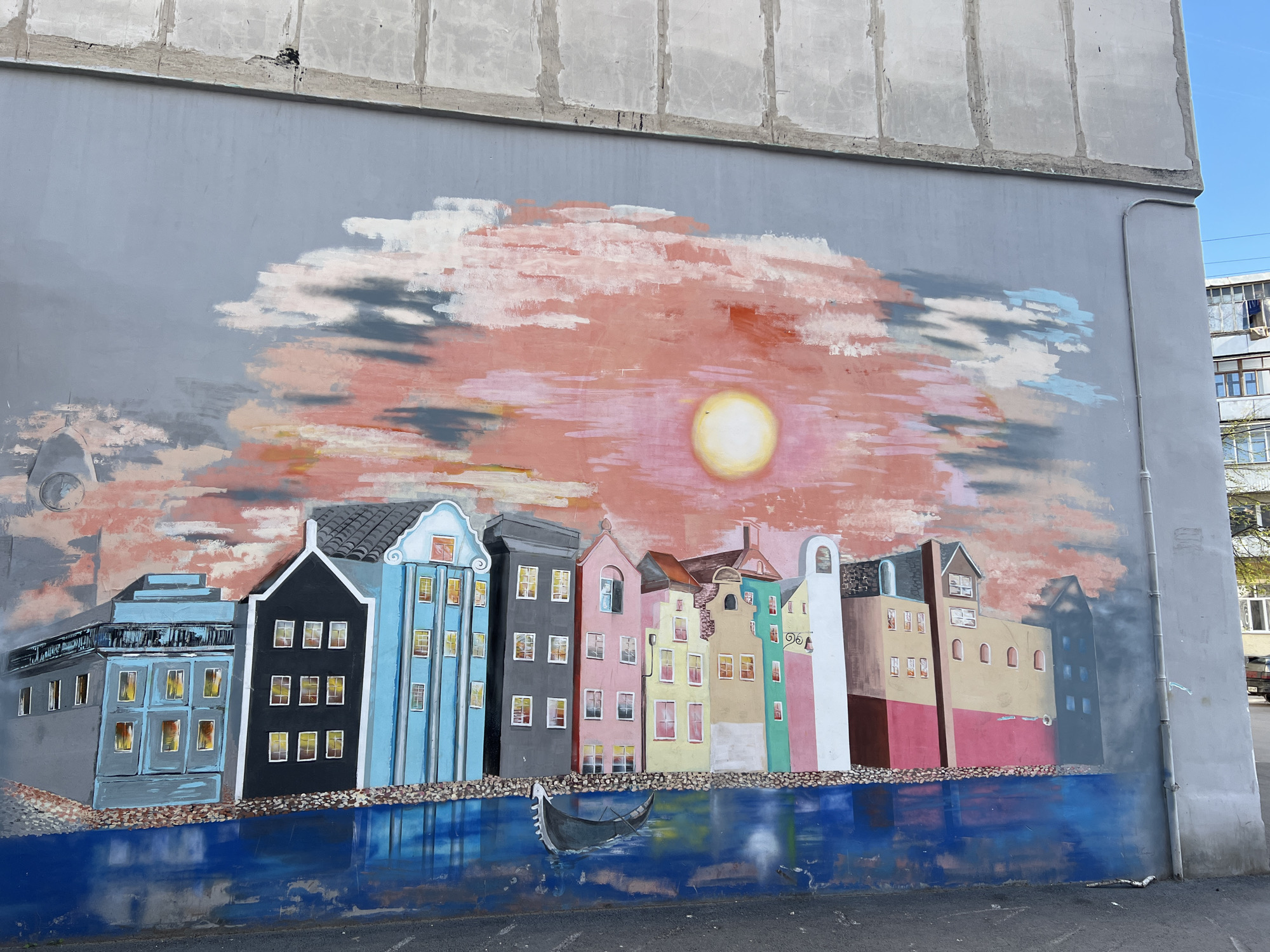 A mural on a grey concrete wall depicts a row of stylized buildings reflected in blue water, reminiscent of Amsterdam or another European canal city. The buildings are painted in various colors, including blue, black, grey, pink, yellow, green, white, tan, and red. Each building has numerous rectangular windows, and the overall style is somewhat cartoonish. In the water is a dark gondola. Above the buildings is a painted sky with a large, pale yellow sun surrounded by pink and grey clouds. The concrete wall extends beyond the mural on both sides, and the top of the wall is visible above the painted sky. The mural appears to be on an exterior wall in an urban environment. Based on the prompt information, the location is Ufa, Russia.