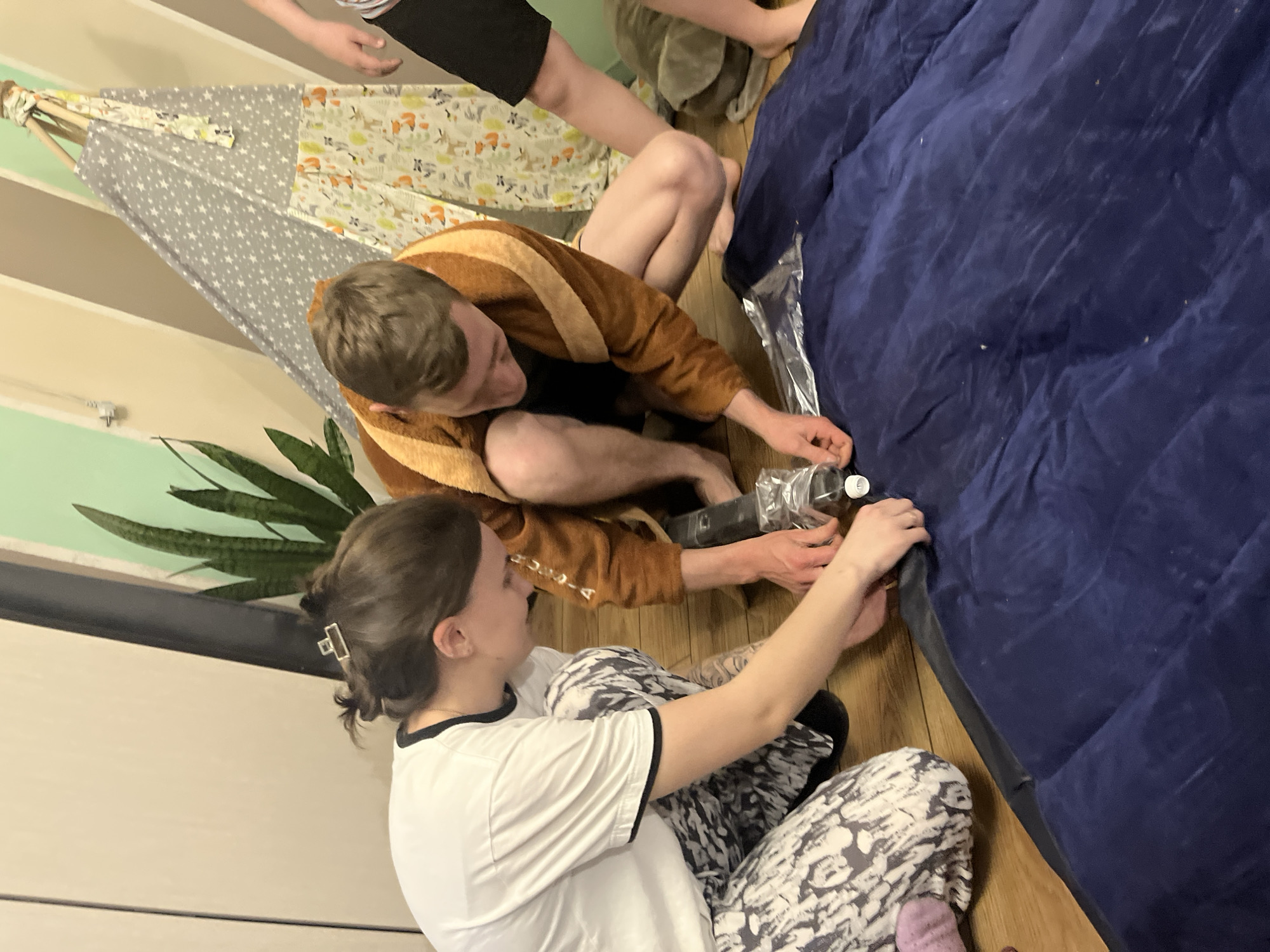 The image shows two individuals inflating a dark blue air mattress on a wooden floor. The person on the left is a woman with dark hair pulled back in a claw clip, wearing a white t-shirt with black trim and patterned pajamas. She is holding the air mattress and the neck of a clear plastic bottle. The person to the right is wearing a brown and yellow striped bathrobe, and they are holding the neck of the bottle near the air mattress's inflation point. In the background, there is a child wearing black shorts and a patterned shirt, and a tent-like structure with a gray and white patterned fabric. A potted plant is visible to the left, and the wall is painted with vertical stripes of beige and mint green. There is no visual evidence to determine the location is Oryol-Izumrud, Russia.
