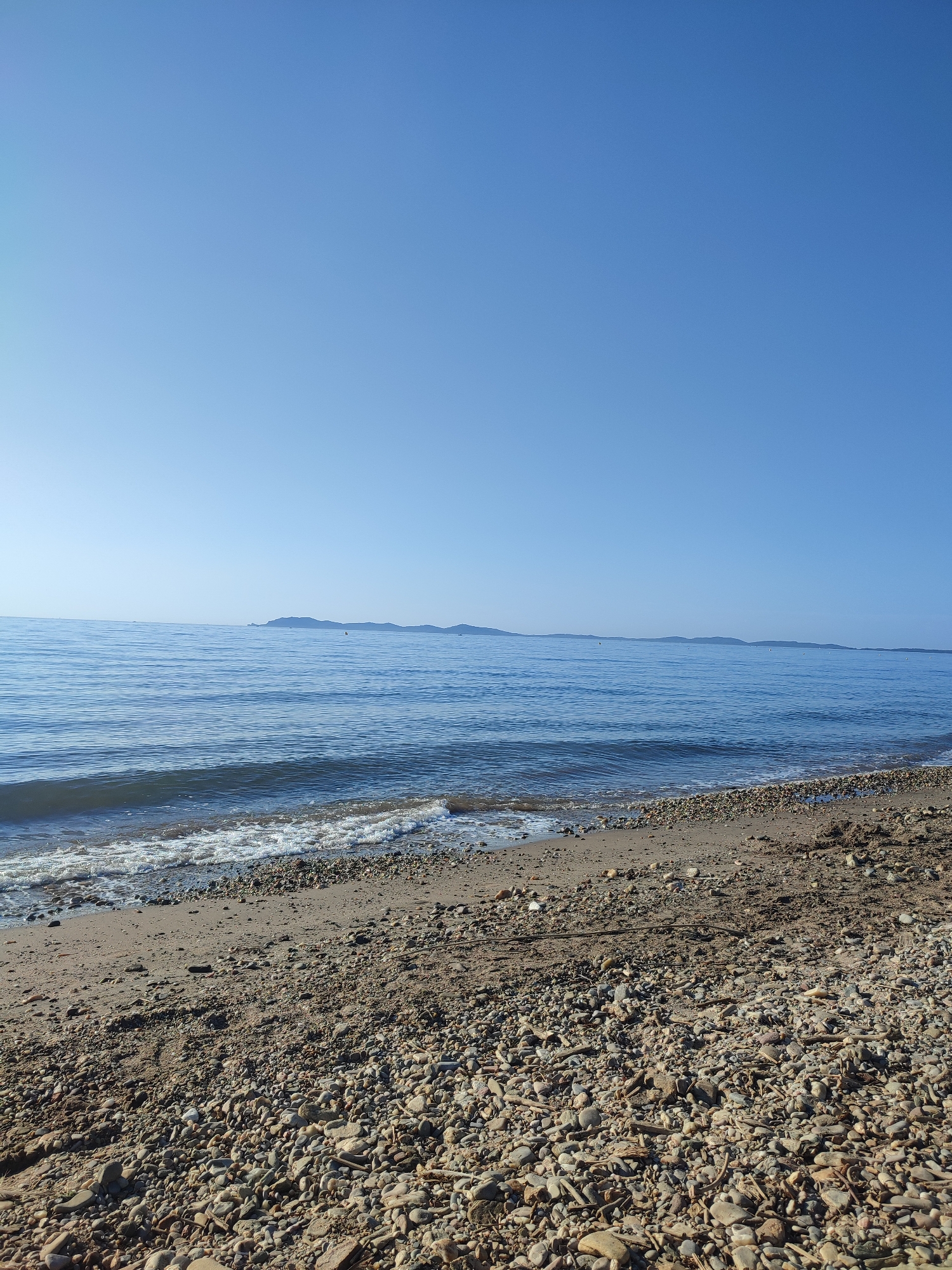 The image depicts a wide, serene view of the Mediterranean Sea from the shoreline of Hyères, France. The foreground is a pebble and sand beach, littered with driftwood and assorted natural debris. Gentle waves lap at the shore, creating a frothy white edge against the darker, wet sand. The sea itself is a calm, deep blue, stretching towards a distant, low-lying coastline characterized by rolling hills or islands. The sky above is a clear, unblemished light blue, indicative of fair weather. There are no people or man-made structures immediately visible in this coastal landscape. The image captures a moment of natural tranquility on the French Riviera.