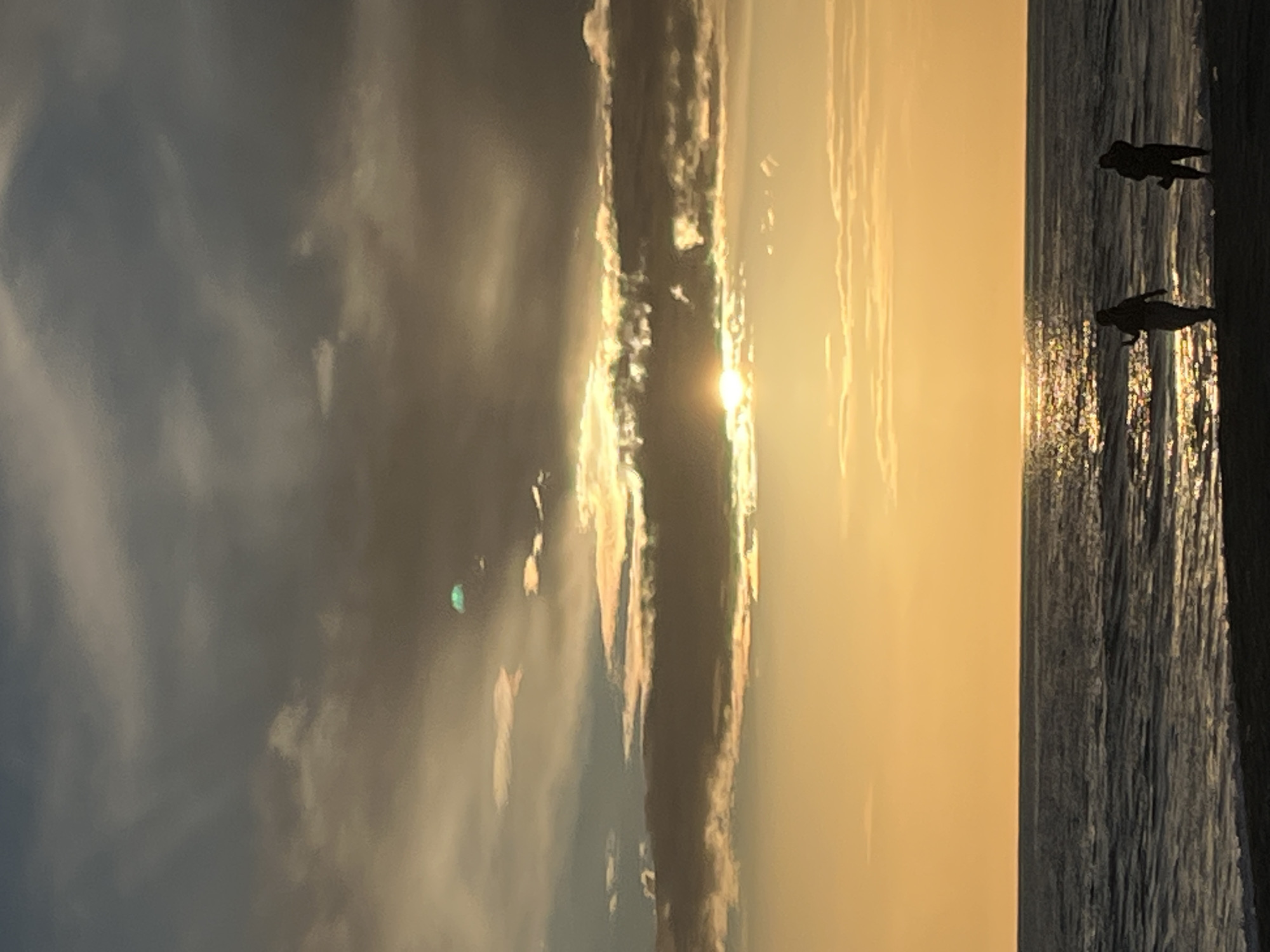 The image depicts a beach scene at sunset, likely in Alanya, Turkey, given the warm, golden light. Two silhouetted figures are visible on the right, standing at the water's edge. The sea reflects the setting sun, creating a shimmering effect. The sky is partially covered by clouds, with the sun's rays breaking through.