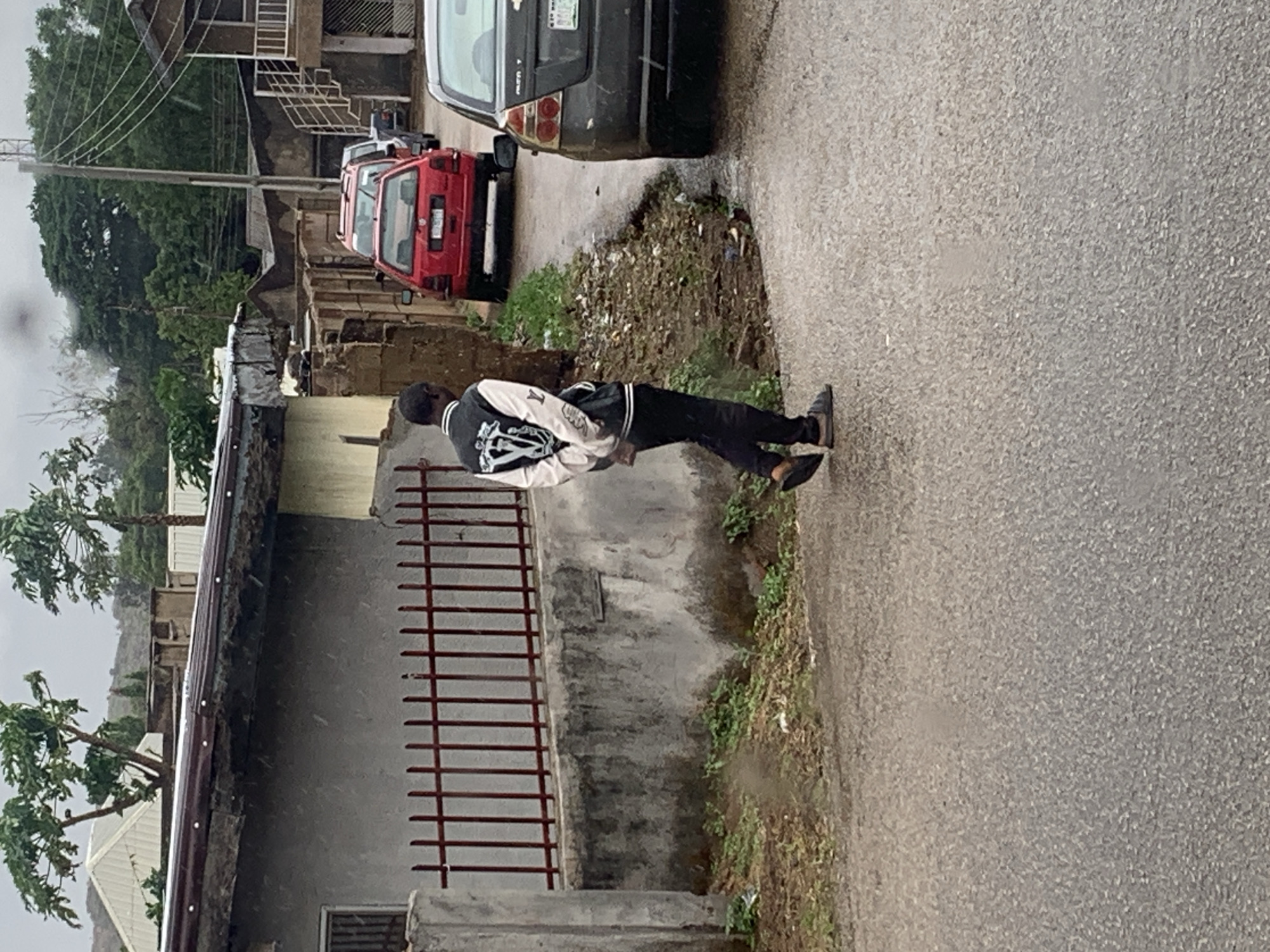 The image depicts a street scene in Jos, Nigeria, on an overcast day, possibly during or after light rain, as the asphalt road appears damp and small white specks resembling raindrops are visible in the air. A person, viewed from behind, stands on the edge of the paved road, wearing a two-toned varsity-style jacket with a large white graphic on the back, dark trousers, and shoes.
Two cars are parked along the roadside: a red hatchback, possibly a Volkswagen, and behind it, the rear of a black sedan. The red car's license plate has "LAGOS" written at the top. To the left of the road, a low concrete wall fronts a dirt embankment with sparse green vegetation. Behind the wall, a building with a grey facade and a reddish-brown metal gate/fence is visible. Further into the background, several other buildings with varied architectural styles and rooflines can be seen, interspersed with lush green trees, including what appear to be papaya trees. A utility pole with power lines stands tall on the left side of the frame, and a metal staircase is visible on one of the distant buildings. The landscape extends to hazy, tree-covered hills in the far background.
