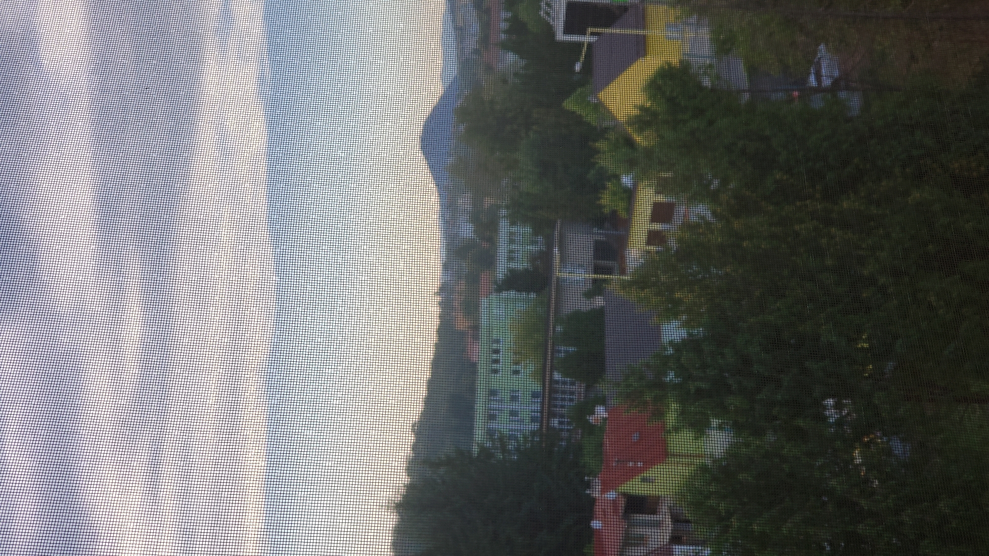 The image depicts an overcast sky above a residential area. In the mid-ground, several multi-story buildings are visible, some painted green and others with a lighter facade. In the foreground, rooftops of houses, primarily red and yellow, are interspersed with trees. A distinct conical hill or mountain is visible in the background, situated to the right of a forested slope. The scene appears to be taken through a window screen. The location is identified as Prešov, Slovakia.
