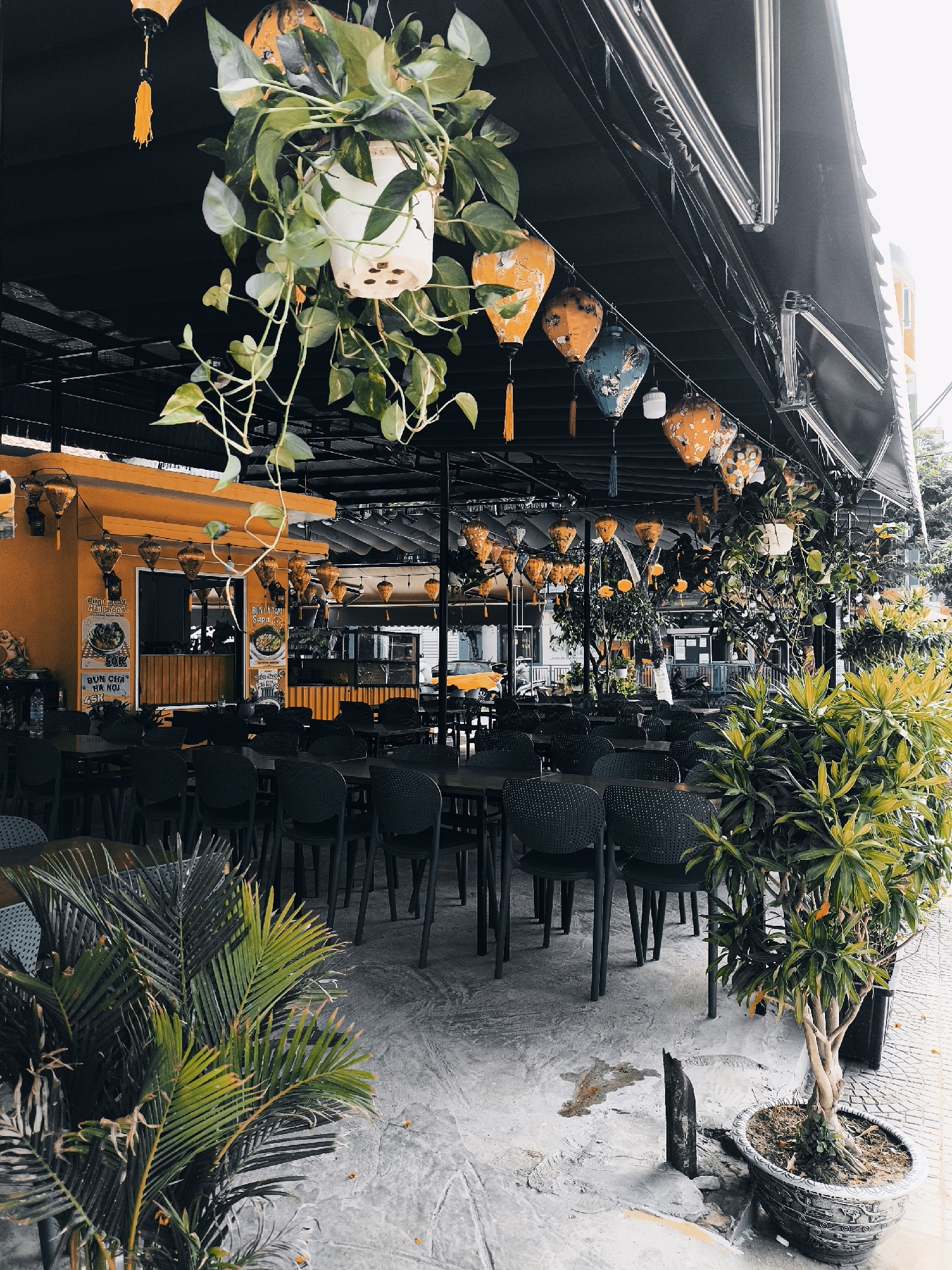 This is a daytime exterior shot of an outdoor dining area of a restaurant in Đà Nẵng, Vietnam. The restaurant has an orange facade and is covered by a black awning. The dining area is furnished with numerous dark-colored chairs and tables. Hanging plants and traditional Vietnamese lanterns, decorated with floral patterns in orange and blue, are suspended from the awning. Potted plants, including a small tree with yellowing leaves and a palm-like plant, are placed around the seating area. The ground is paved with concrete and some decorative tiles. The image appears to have a slightly desaturated and high-contrast aesthetic. No people are visible in the scene.