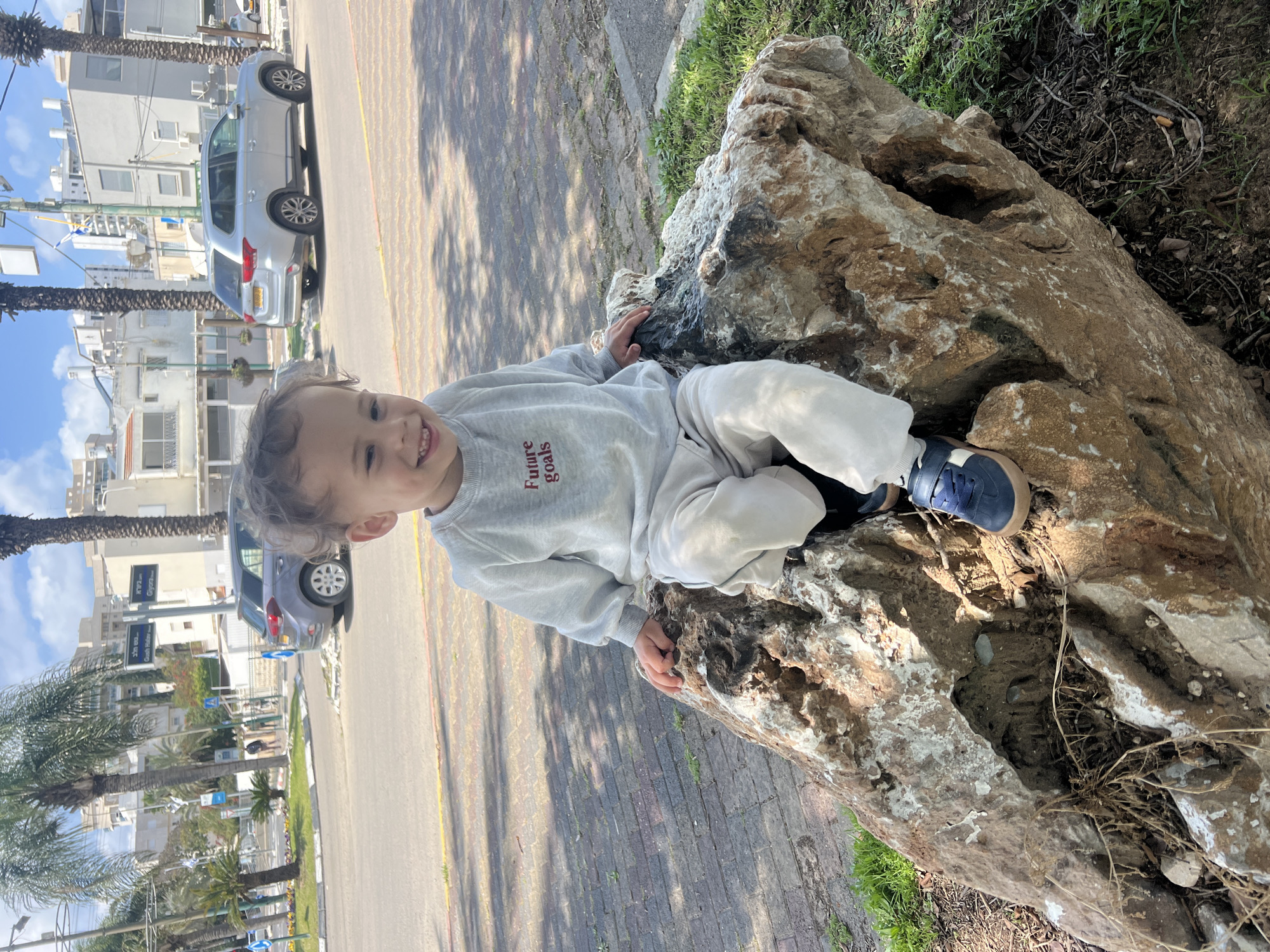 A young child is sitting atop a large rock formation in an outdoor setting. The child, dressed in a grey sweatshirt with "future goals" written on it in red letters, and light-colored pants and blue sneakers, is smiling directly at the camera.
The rock formation is brown and tan with white streaks, and appears to be part of a natural area with some grass and fallen debris nearby. Beside the rock and to the left, there is a paved area comprised of brown brick and asphalt, with shadows cast on the surface. A street runs along the left side of the frame, featuring parked cars, palm trees, and buildings. Street signs in Hebrew are also visible.
The photograph was taken in Kiryat Bialik, Israel.