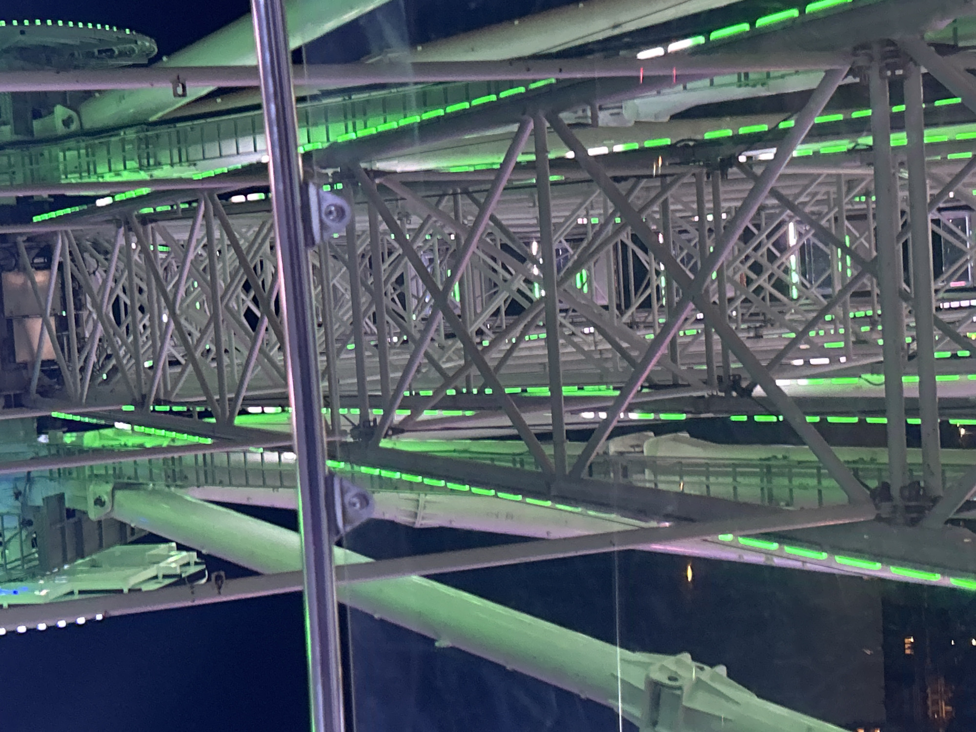 This is a close-up, low-angle, vertical, exterior shot of a Ferris wheel at night, likely located in Szczecin, Poland. The structure of the Ferris wheel is made of a complex network of grey metal beams, illuminated by numerous green LED lights that line the edges of the structural elements. The sky is dark, indicating it is nighttime. In the foreground, a horizontal metal railing with a reflective surface is visible, suggesting the shot is taken from a viewing platform or another nearby structure. The green lights create a striking contrast against the dark background and the metal framework, highlighting the intricate geometric patterns of the Ferris wheel's construction. The overall impression is one of modern urban infrastructure with decorative lighting.