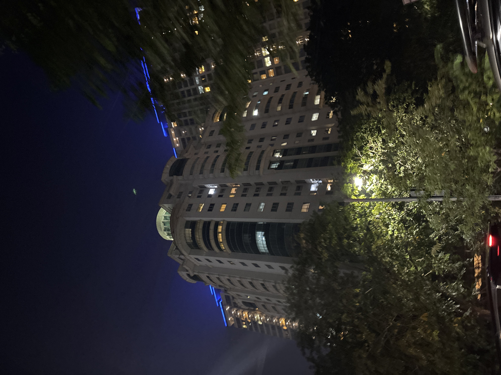 A tall apartment building is illuminated at night in South Jakarta, Indonesia, with blue lights along its rooftop and lights visible through its windows. Trees with green leaves obscure the lower portion of the building, and some of the trees are lit by a streetlamp. The sky is dark blue.