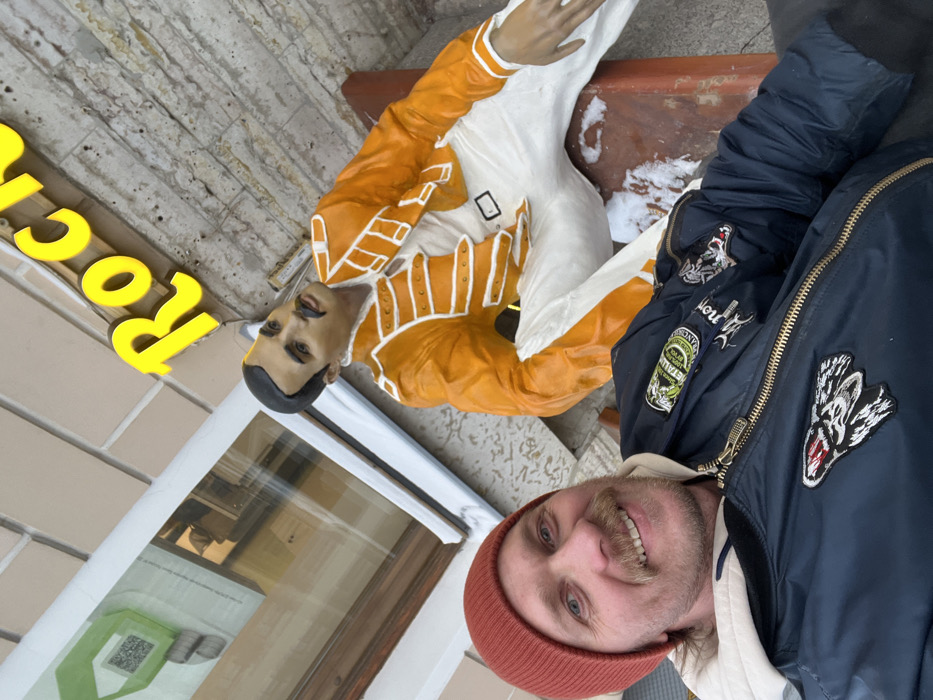 An outdoor photograph captures a smiling male subject positioned alongside a life-sized statue of Freddie Mercury. The scene is located in Saint Petersburg, Russia, outside the Rock Cafe at Dumskaya Street 1/3, indicated by the visible yellow neon sign partially reading "Roc..." and the characteristic statue.
The male subject, positioned in the lower left foreground, presents a direct, smiling gaze at the camera. He has blonde hair partially visible beneath an orange knit beanie, and a prominent blonde mustache. He wears a light-colored hooded sweatshirt under a dark blue bomber jacket, which features embroidered patches including a white wolf's head and a 'Metallica' logo.
To the right and slightly behind the male, the statue of Freddie Mercury is depicted in a seated position on a reddish-brown bench. The statue wears an iconic orange military-style jacket with white detailing, white trousers, and a black belt. It features a mustache. Patches of snow are present on the bench surface adjacent to the statue's right leg. The background includes a light-colored brick wall and a window with a white frame, reflecting an interior light source and a green graphic.