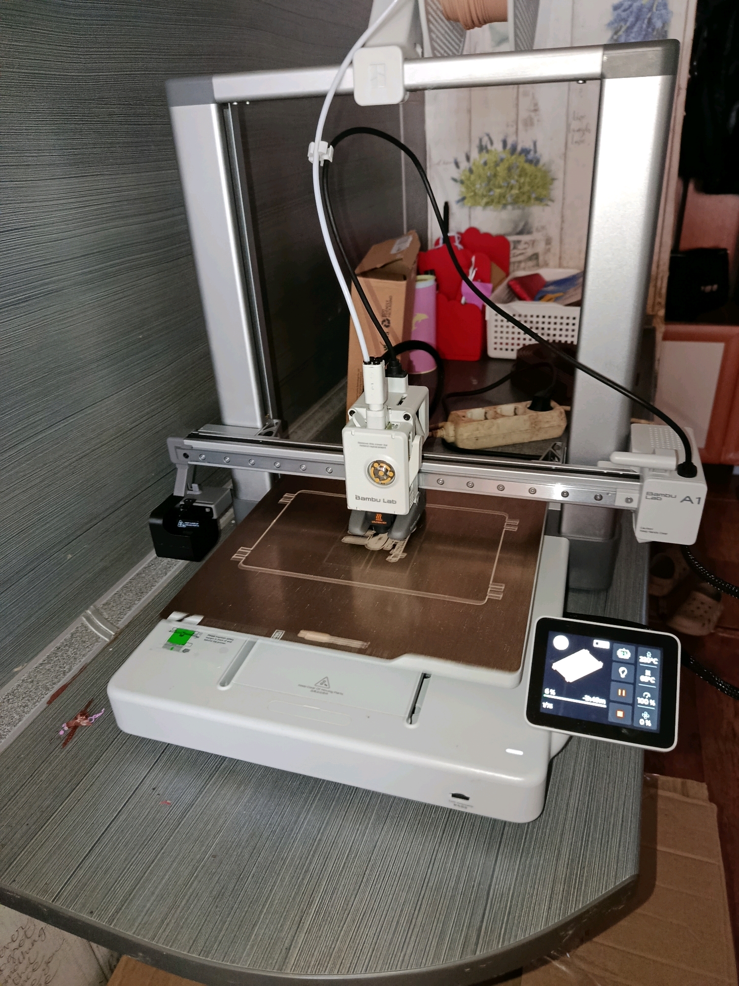 The image shows a 3D printer, specifically a Bambu Lab A1, situated indoors. The printer is white and silver, with a build plate and print head visible. A small touchscreen interface is attached to the printer, displaying various icons and data points related to the printing process. The background suggests a home environment, with shelves and boxes visible. The textured gray wall and wooden furniture indicate a domestic setting, possibly in a room within a dwelling. The presence of a power strip suggests the printer is connected to an electrical source. The overall scene depicts a functional 3D printing setup in a residential space. The location in the city of Fokino, Russia, is not discernible from the visual information provided in the image.