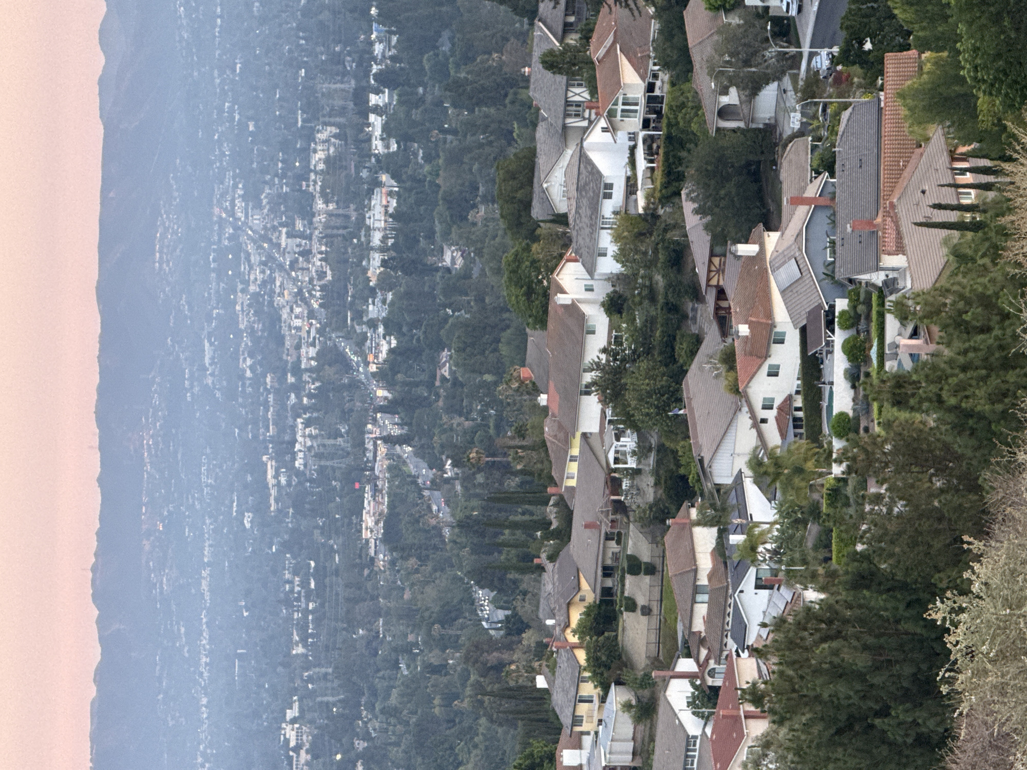 The image captures a panoramic view of a residential neighborhood nestled in the hills of Los Angeles, California, USA. The scene is illuminated by the soft, warm light of either dawn or dusk, casting a gentle pink hue across the sky and silhouetting the distant mountain range. The foreground is dominated by a cluster of single-family homes, their varied architectural styles and rooflines creating a sense of suburban sprawl. These houses are interspersed with lush greenery, including mature trees and manicured shrubs, suggesting a well-established community.
As the eye moves towards the middle ground, the density of the housing increases, with more rooftops and structures visible. A road can be seen winding through the neighborhood, with a few vehicles present, indicating ongoing activity. The background opens up to a wider cityscape, characterized by a hazy, atmospheric perspective. Numerous buildings, lights, and infrastructure elements are discernible, hinting at the vastness of the Los Angeles urban landscape. The overall impression is one of a sprawling, affluent residential area situated within a larger metropolitan context. There are no visible people or specific actions taking place, rendering the scene a static, observational snapshot of the environment.