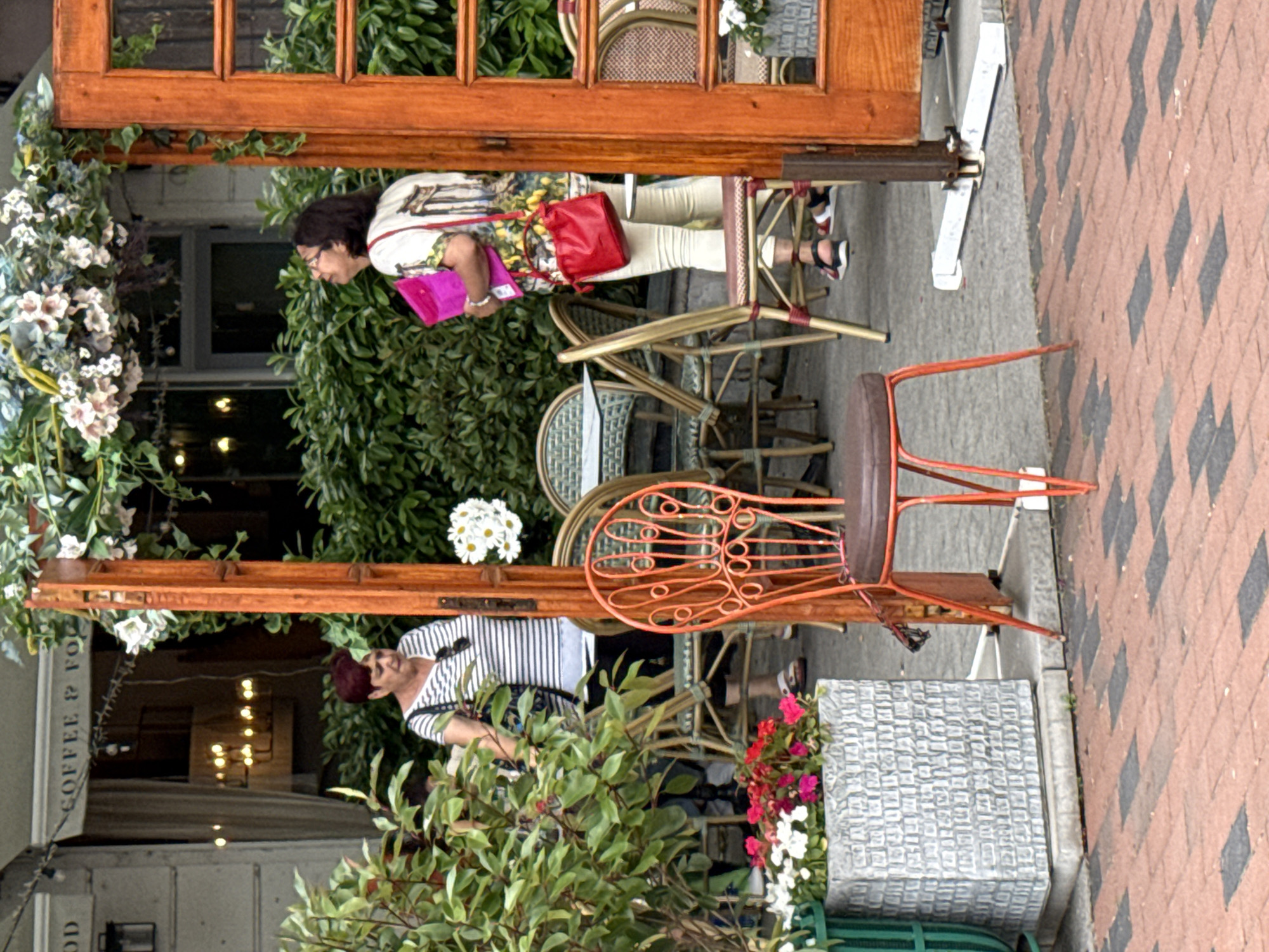 A medium shot shows an outdoor seating area at a cafe. A woman with dark hair and glasses stands looking up and to the left, holding a bright pink folder and a red purse. She wears a white top with a colorful floral pattern and light-colored pants. To her left, another woman with short, dark red hair and a striped shirt looks down. She carries a dark bag on her shoulder. In the foreground, an orange metal chair with a brown seat is positioned to the right of a grey planter filled with red and white flowers. Behind the women, there are tables and chairs with wicker backs and seats. A wooden door frame with a glass pane is on the right side of the image, with green foliage cascading down. The ground is paved with red and black bricks. The background consists of a building with a sign that reads "COFFEE & FO" and dense green bushes. The scene appears to be a street-level cafe.