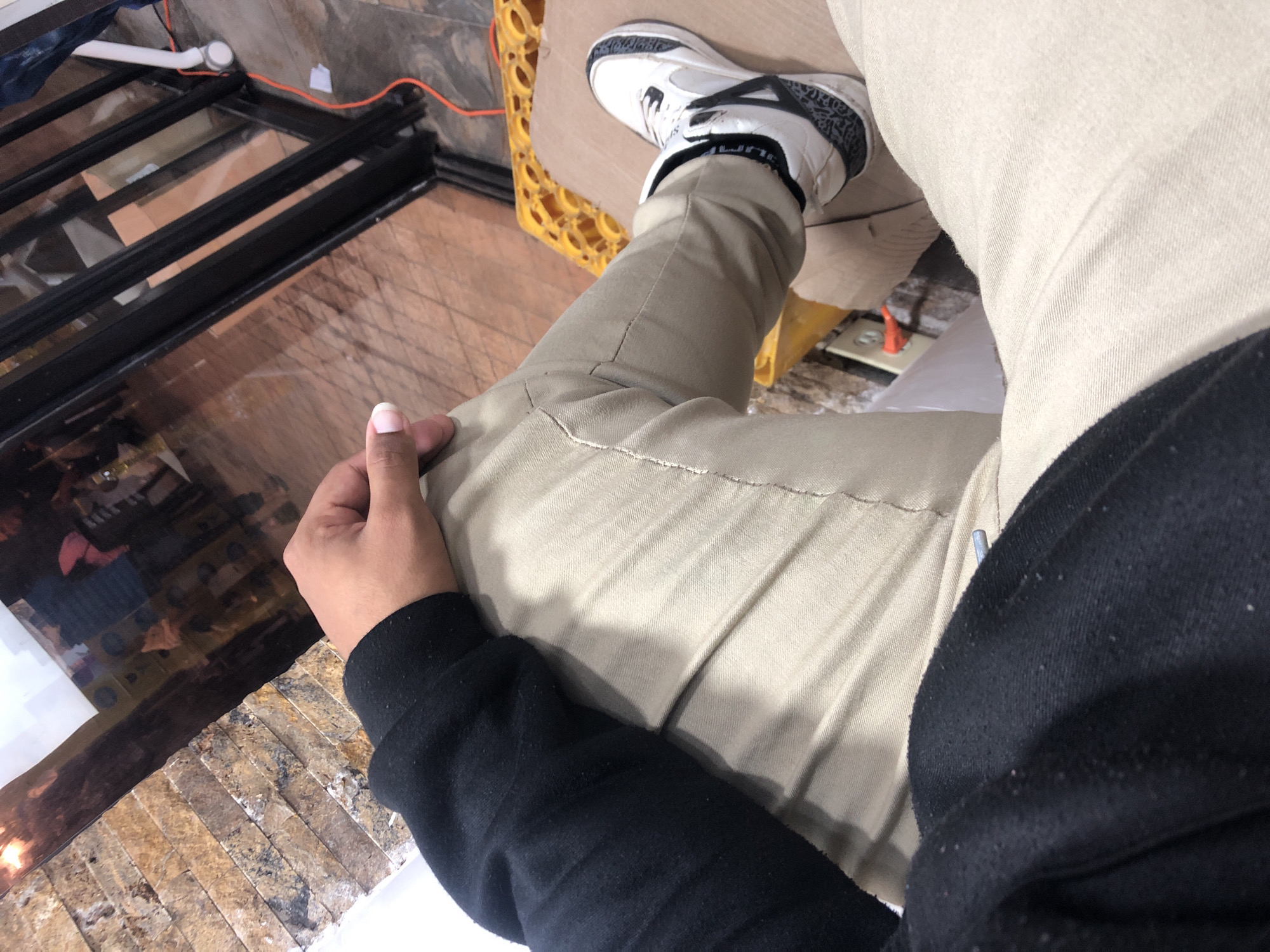 The image, taken from a high-angle perspective, captures a person's lower body and right arm in what appears to be an indoor or semi-indoor setting in Juan Griego, Venezuela. The person is wearing light khaki or beige pants and a black long-sleeved top or sweatshirt, which shows some pilling on the fabric. On their right foot is a white sneaker with black accents and a distinctive elephant print design, characteristic of an Air Jordan 3 shoe; the word "JUMPMAN" is visible on the sock or ankle cuff. The foot is resting on a yellow or orange object, possibly a crate or a support structure.
The person's right hand, with light pink painted fingernails on the index finger and thumb, rests on their left thigh. In the background to the left, a large reflective surface, likely glass, shows indistinct reflections of what appears to be an interior space with furniture or other objects. Below this reflective surface is a textured wall or counter made of stacked, light-colored stone or tile.
The floor in the top right area of the image is a plain, possibly tiled or concrete surface, appearing somewhat raw or unfinished. An orange electrical cord snakes across this floor, leading to a white pipe or conduit and an electrical outlet visible on the wall or floor. The overall impression is a blend of utilitarian elements (exposed wiring, pipes) and more finished architectural details (stone, reflective glass), suggesting a construction phase, renovation, or a functional space.
