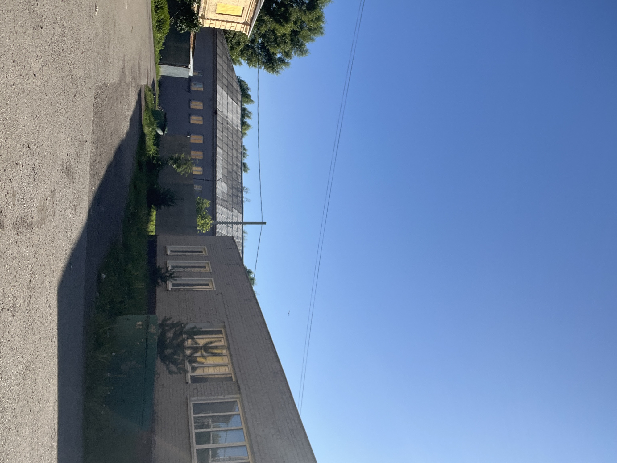 The image depicts a bright, sunny day in a semi-industrial area, likely in Kiselyovsk, Russia. The scene is dominated by a clear blue sky, with a few thin power lines stretching across it. In the foreground, a grey asphalt road curves into the frame from the left, casting a long shadow from an unseen object.
To the right of the road, a row of buildings with dark grey facades is visible. The buildings have numerous rectangular windows, many of which appear to be illuminated from within or reflect the sunlight with a yellowish hue. The windows are arranged in regular patterns along the multi-story structures. One building, in the foreground, is constructed of grey brick. A large, dark green structure, possibly a storage container or equipment housing, is situated near the base of this building.
In the background, partially obscured by trees and other buildings, is another structure with a lighter facade, featuring rows of rectangular windows. Lush green trees with dense foliage are interspersed throughout the scene, suggesting a temperate climate. The overall impression is one of a typical working day in a city or town, with clear weather and good visibility. There is no visible text in the image.