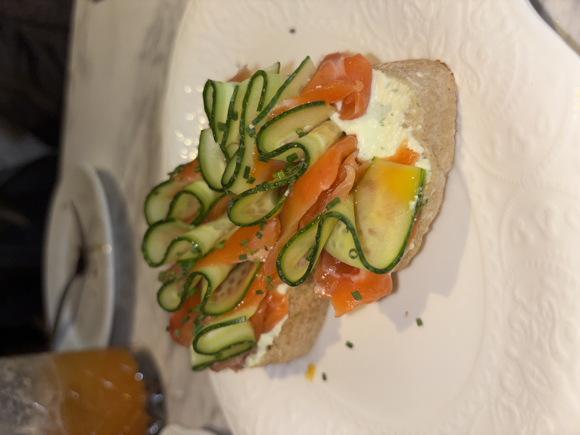 A close-up, angled overhead shot shows two open-faced sandwiches on a white plate with a subtle embossed pattern. Each sandwich is constructed on a slice of brown bread and topped with a creamy spread, likely cream cheese or a similar dairy product. Slices of salmon and spiraled cucumber ribbons are layered on top. Chopped chives are sprinkled as a garnish. The sandwiches are placed on the right side of the plate. To the left and out of focus are parts of a table setting that include a stack of white dishes and an orange-brown object encased in clear plastic. The environment appears to be an indoor setting, possibly a restaurant. There is no indication to confirm the location to be Saint Petersburg, Russia.