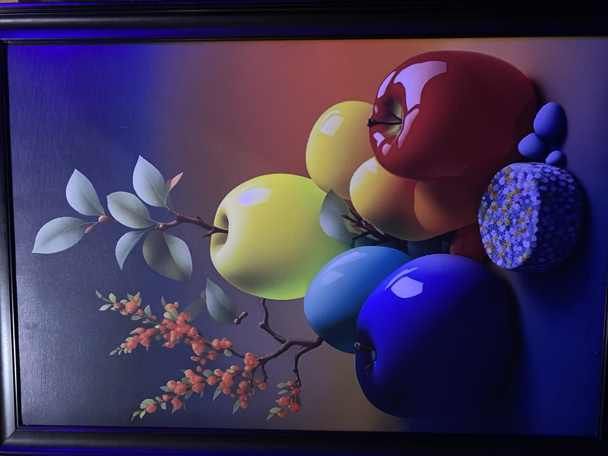 An image displays a framed, modern still-life artwork, likely photographed indoors under low light conditions with a distinct blue light source illuminating the top left. The artwork features an arrangement of highly stylized, glossy apples and botanical elements against a gradient background that shifts from deep blue on the left to warm reddish-orange on the right.
Prominent objects in the artwork include several vibrantly colored apples: a large, shiny red apple at the top right, multiple bright yellow apples, a light blue apple, and a dark blue apple at the bottom right. The apples have a smooth, almost plastic-like finish, reflecting light strongly. Interspersed among the apples are tree branches adorned with pale green leaves and clusters of small, reddish-orange berries or flowers. On the far right, a cylindrical or half-sphere object with a white and yellow floral pattern against a purple background is visible, alongside a few small, smooth, blue, pebble-like shapes.
The composition suggests a whimsical or surreal take on traditional still life. No activity or event is taking place; it is a static display. The ambient blue lighting visible on the frame and parts of the background of the image is a notable detail of the photograph itself, not the artwork. There is no visible text in the image. While the image was captured in Badagry, Nigeria, the artwork itself does not contain any geographical cues to this location.