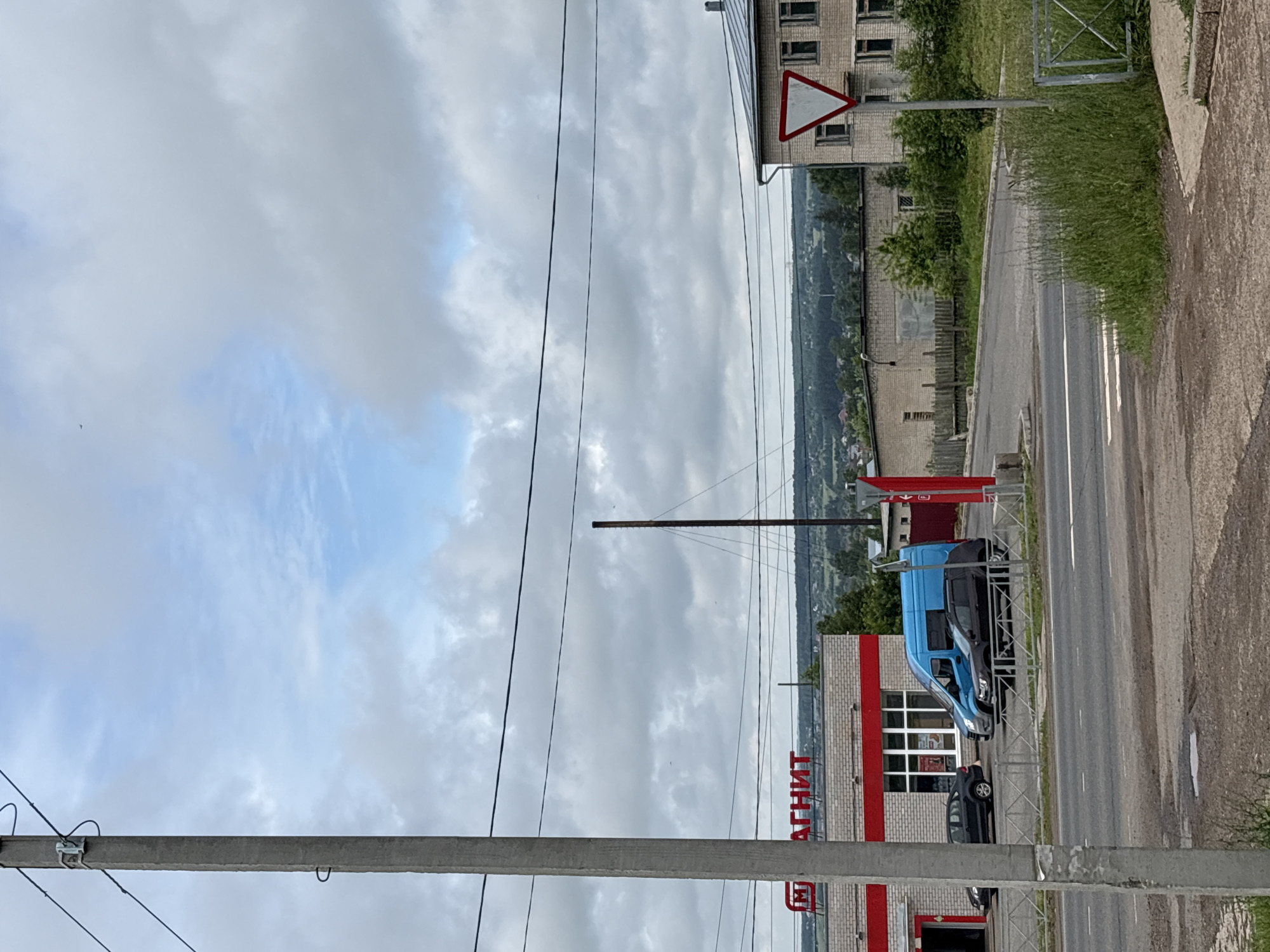 The image captures a street scene in Kstinin, Russia, on a cloudy day. In the foreground, a concrete utility pole stands to the left, with overhead wires. A paved road with a pedestrian crossing is visible. To the right, a patch of tall grass and a metal fence line the sidewalk. In the mid-ground, a red and white building with the Cyrillic lettering "МАГНИТ" (Magnit) is prominent, suggesting a retail store. Several vehicles, including a blue van and two dark-colored cars, are parked in front of the store. A red post stands near the entrance. Further in the background, a multi-story brick building with numerous windows is visible, along with a triangular yield sign on a pole. The horizon is lined with trees and buildings, indicating a rural or semi-urban landscape. The sky is overcast with scattered clouds. There are no people clearly visible in the image.