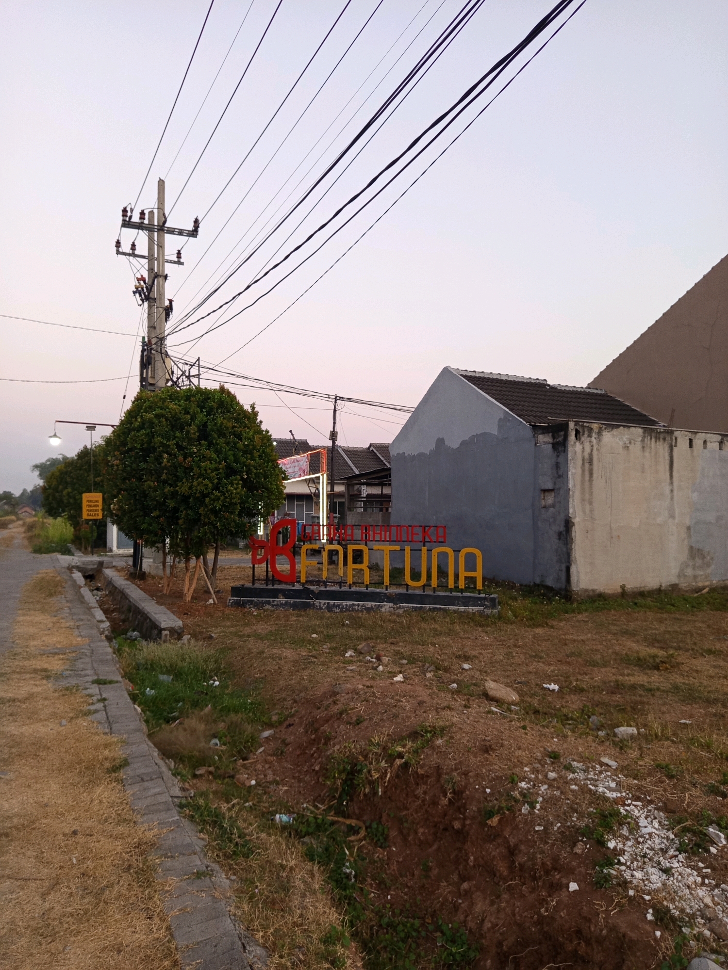 The image captures a street scene in Ngastemi, Indonesia, likely at dusk or dawn given the soft lighting. A concrete road with dry grass on the side runs diagonally from the lower left corner. Adjacent to the road is a concrete drainage ditch. In the mid-ground, a prominent power pole with multiple wires stretches upwards against a pale sky. Two rounded, manicured green trees stand near the power pole. In front of the trees, a red and yellow sign spells out "B FOR FORTUNA." Behind the sign and trees, a grey and beige building with a tiled roof is visible, appearing somewhat weathered. Further in the background, other buildings and more power lines can be seen. The overall environment is somewhat unkempt, with dry vegetation and some scattered litter in the ditch and surrounding ground. No people are visible in the frame.