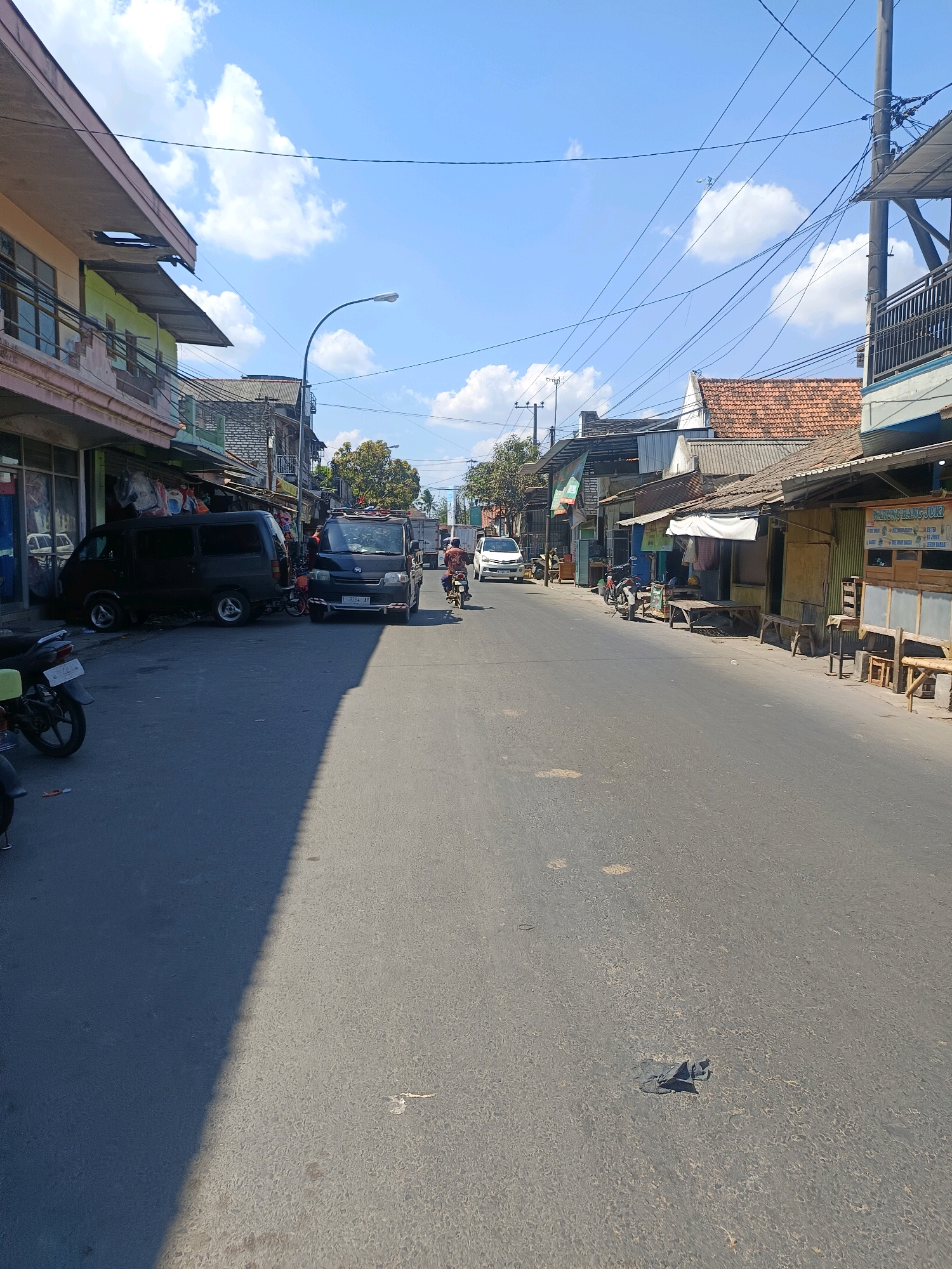 The image depicts a street scene in the city of Blega, Indonesia, under a bright, sunny sky with scattered clouds. The perspective is from the middle of the paved road, looking down its length.
On the left side of the road, there are several storefronts with awnings. A dark-colored van is parked on the left side, partially obstructing the view of the storefronts. A motorcycle is parked in front of one of the shops.
On the right side of the road, there are more shops, some with wooden benches and tables outside. One shop has a sign that reads "WARUNG BANG JUki" and lists various drink options. There are also several wooden structures that appear to be part of the shops.
The road itself is asphalt and shows some signs of wear. There are shadows cast across the road from the buildings and a large shadow dominates the foreground on the left side, suggesting the sun is high in the sky.
Several vehicles are present. Two dark-colored vans are parked on the left side. A white car is seen driving away from the camera on the left. A motorcycle with a rider is proceeding down the road on the right side. Another motorcycle is parked on the left.
Power lines and utility poles are visible crisscrossing the sky above the buildings, a common feature in many urban and semi-urban environments. The overall impression is of a bustling, though not overly crowded, street scene in a developing urban area.