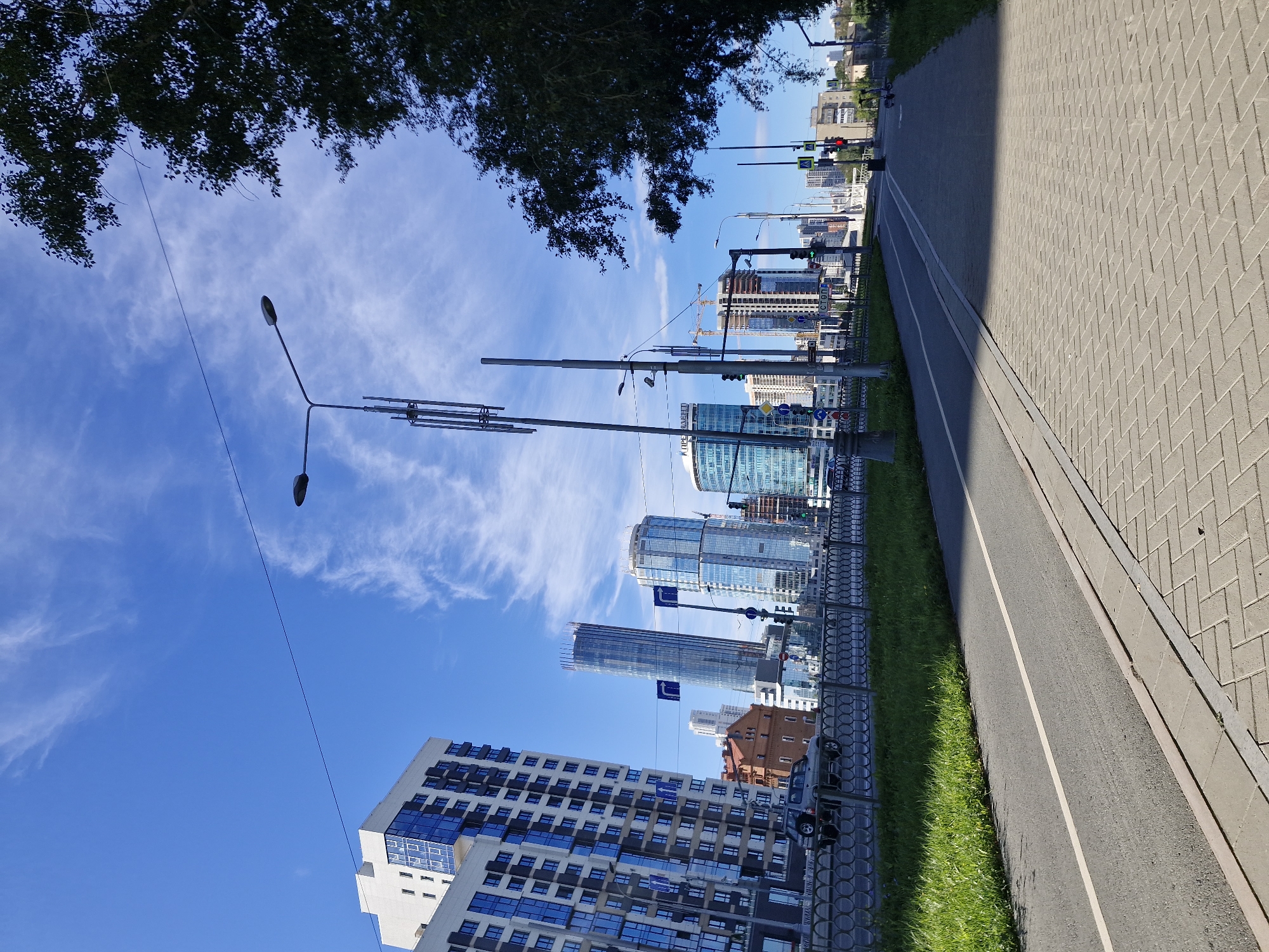 The image depicts a street scene in Yekaterinburg, Russia, likely in the vicinity of the Iset River embankment or a similar urban area with modern architecture. The foreground features a paved pedestrian walkway with a brick pattern and an adjacent asphalt bike lane marked with a white line. To the left of the walkway is a strip of green grass and then a road with a silver SUV parked on it. Beyond the road, a metal decorative fence separates it from the green space.
The background is dominated by a clear blue sky with thin, wispy white clouds. Several modern high-rise buildings are visible, characterized by their glass facades and contemporary designs. These include a tall, slender cylindrical building and a wider building with blue-tinted windows. Other structures, including a red brick building and buildings under construction with cranes, are also present. Streetlights and traffic signals are visible, suggesting an active urban environment. A large tree with dark green leaves shades a portion of the right side of the image. The scene appears to be captured on a sunny day.