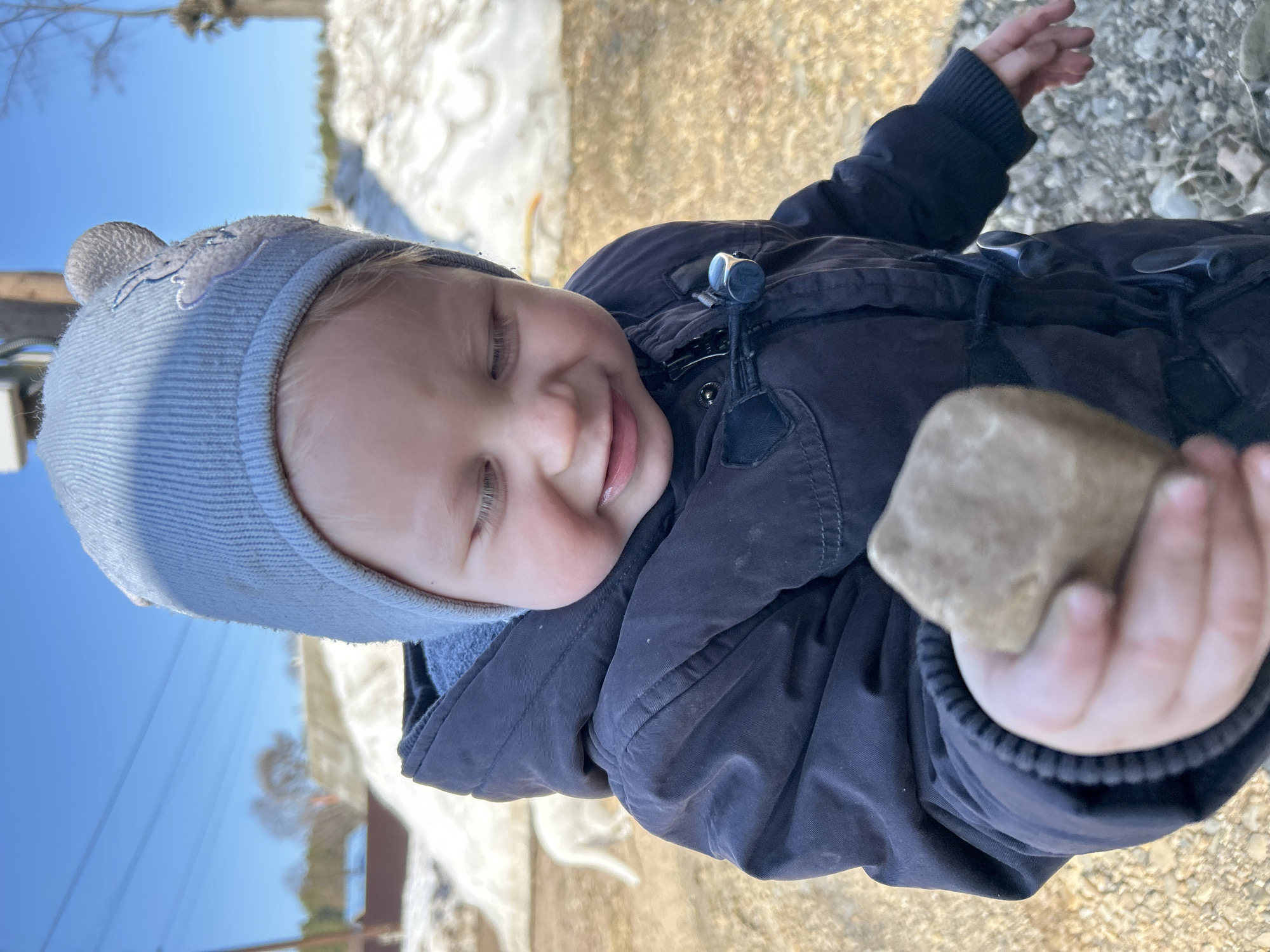 A young child in a blue hat and dark jacket is holding a rock. The child's eyes are partially closed, and they appear to be smiling faintly. The background shows patches of snow and a clear blue sky. The location is outdoors, possibly near a park or natural area in the city of Chaikovsky, Russia.