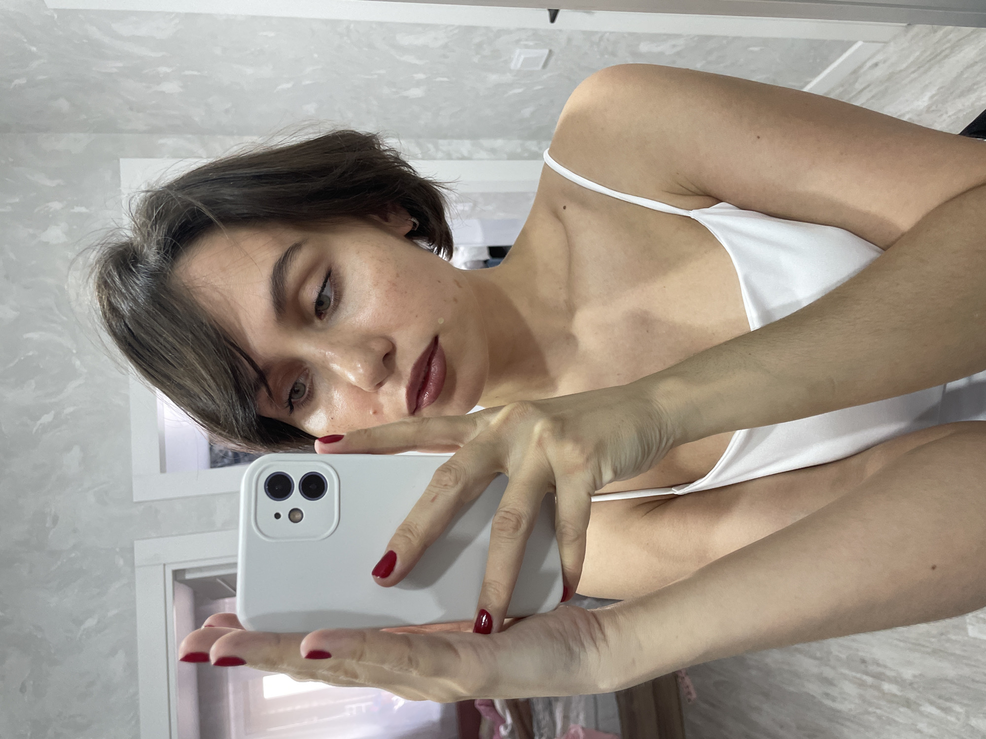 A woman with short brown hair and red nails is taking a selfie in a room with light grey marbled walls. She is wearing a white spaghetti-strap top. The phone in her hands has a white case. A partially visible doorway is in the background. It is not possible to determine if the location is Izhevsk, Russia.
