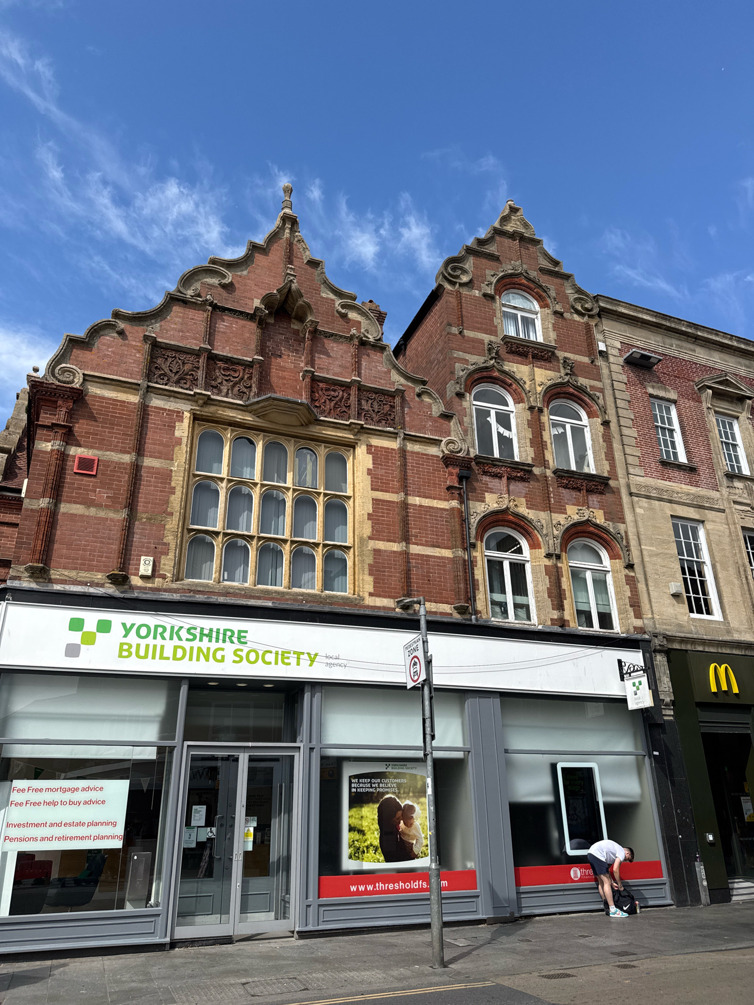 The image shows the exterior of the Yorkshire Building Society on a street in Exeter, United Kingdom. The building features red brick with decorative stonework and arched windows on the upper floors. The ground floor has a large storefront with the Yorkshire Building Society logo prominently displayed. Several signs inside and outside the building advertise financial services. A person is seen bending down on the sidewalk in front of the building, near a McDonald's restaurant which is visible to the right. The sky is blue with some white clouds.