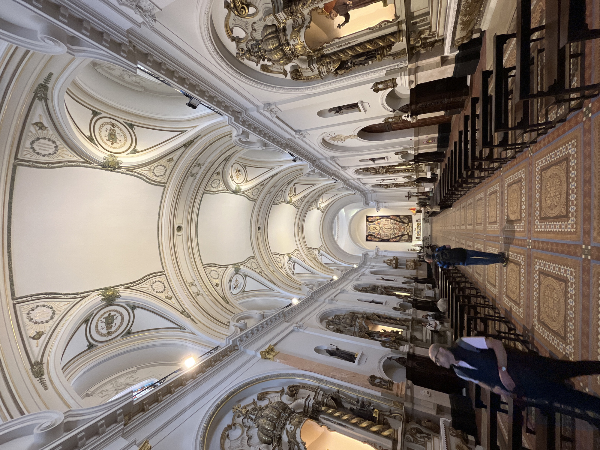 The image displays the grand interior of a church or basilica, viewed from an elevated perspective looking down the long nave towards the altar area. The setting is characterized by elaborate, ornate architecture, typical of Baroque or Neoclassical styles found in historic buildings, consistent with the location context of Buenos Aires, Argentina.
The dominant feature is the intricately decorated vaulted ceiling, adorned with white stucco, gold accents, and grey detailing. Large arches span the nave, each bay featuring elaborate medallions with subtle symbolic or decorative motifs and ornate borders. Recessed lighting fixtures are visible along the upper sections of the walls and arches, illuminating the space.
Along the right side of the nave, rows of dark wooden church pews extend into the distance. The floor is covered in highly decorative patterned tiles, featuring geometric and circular designs in shades of brown, tan, and cream, creating a rich visual texture. The walls are lined with arched alcoves, many of which contain dark statues or religious figures, framed by the same elaborate gold and white ornamentation seen on the ceiling. At the far end of the nave, a large tapestry or artwork, possibly an altarpiece, is visible.
Two individuals are prominently featured in the aisle. In the foreground, a man with a bald or shaved head, wearing a white t-shirt, a dark vest, and dark pants, is seen walking. He has a watch on his left wrist. Further back, another person wearing a blue and black striped shirt and a backpack is standing. Other blurred figures are visible seated within the pews. The lighting appears to be a combination of natural light and artificial illumination from the fixtures, creating a quiet and reverent atmosphere. No legible text is visible within the scene.