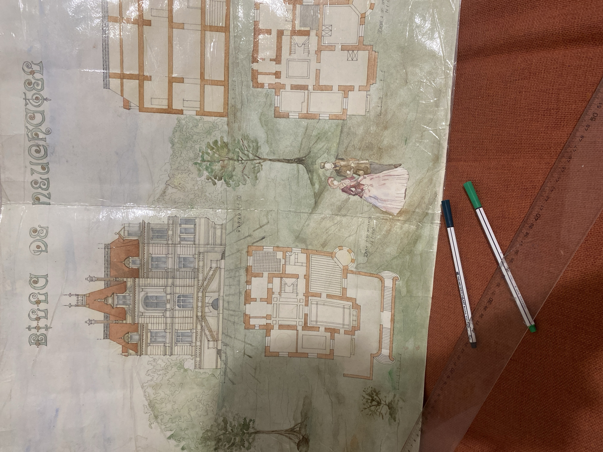 The image depicts an architectural drawing, likely a plan for a building or a complex of buildings, located in Yekaterinburg, Russia. The drawing includes both exterior elevations of buildings and floor plans. One building appears to be a multi-story mansion with detailed architectural features. There's a depiction of people near a tree, suggesting the building is set in a landscape. The text on the drawing includes scales such as "1:00" and "1:100" and the words "Фасад" (Facade) and "План" (Plan) with additional Russian text describing a floor. Also visible are drawing tools: two pens (green and blue) and a ruler. The drawing is seemingly protected under a transparent layer.