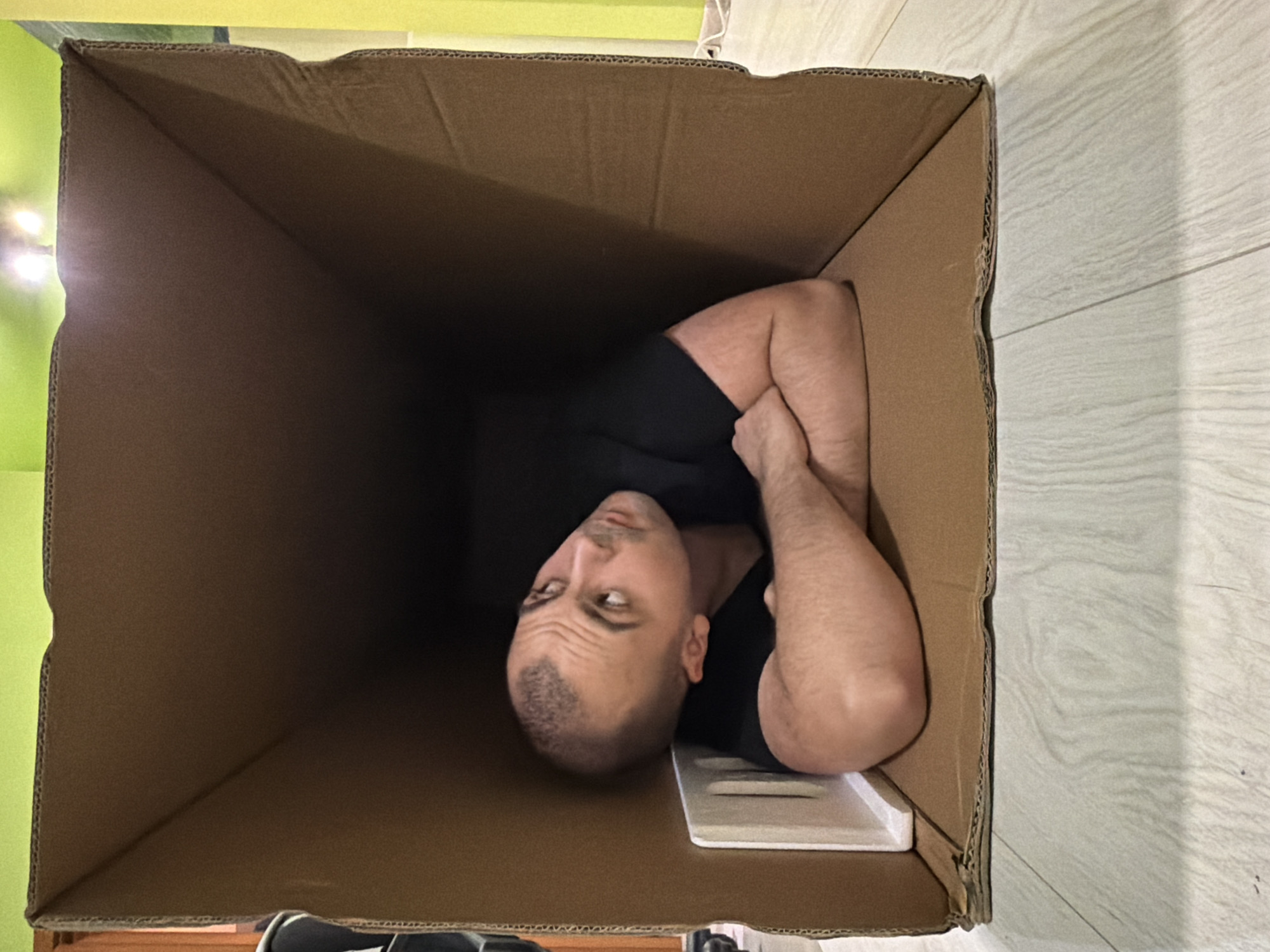 A man is crouched inside a brown cardboard box. The box is situated on a floor that appears to be made of light-colored wood planks. The man is wearing a black t-shirt and has his arms crossed. He is looking up and to his left. A white, rectangular object lies beneath his head. The location is not discernible from the image, so the city of Plovdiv, Bulgaria cannot be verified.