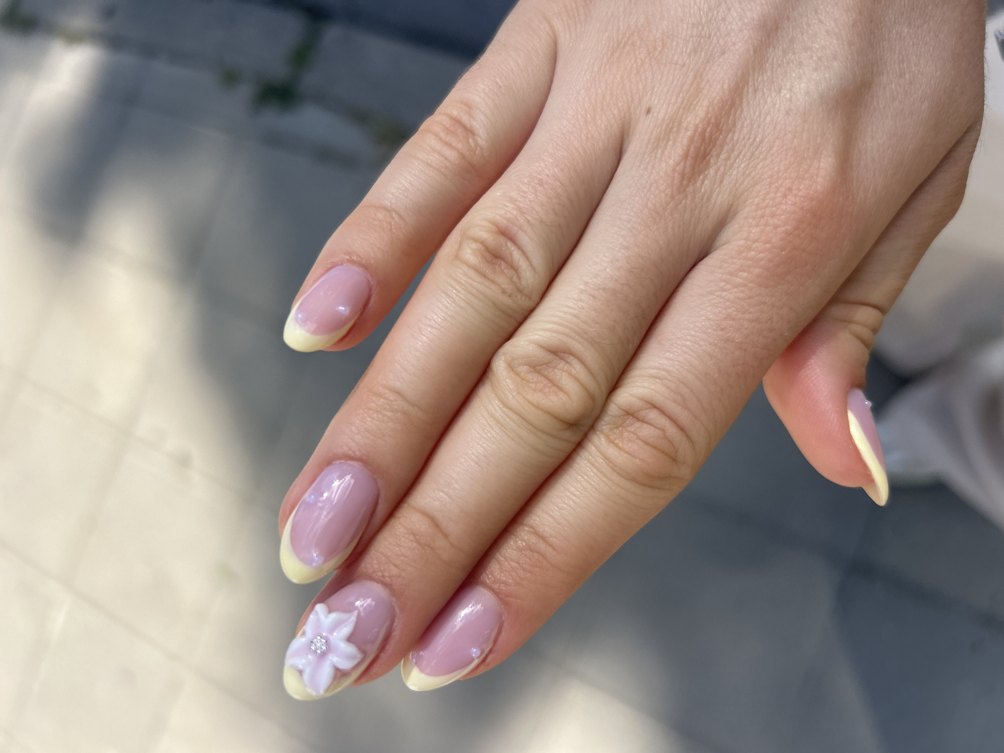 The image shows a close-up view of a person's hand with manicured nails. The nails are shaped into a rounded oval and feature a French manicure design with a sheer pink base and a pale yellow tip. Several nails have small, iridescent embellishments, and the thumb nail has a delicate white flower appliqué with a tiny gem in the center. The hand is positioned over a blurred background that appears to be an outdoor setting with a textured surface, possibly pavement or tiles, illuminated by sunlight. The lighting suggests it is daytime, and the overall mood is serene and aesthetic, focusing on the detailed nail art. No visible text is present in the image.