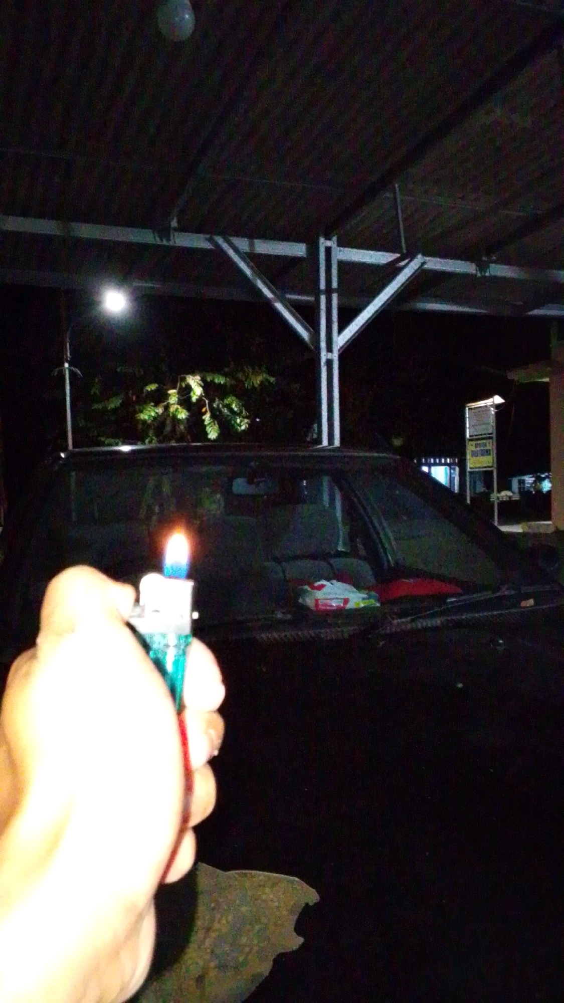 A person's hand is holding a lit teal disposable lighter in the foreground. The lighter is positioned in front of a dark-colored car with its hood and windshield visible. Inside the car, on the dashboard, are a pack of tissues and a red object, possibly a cloth or a bag. In the background, a street light illuminates trees and foliage. The scene appears to be outdoors, likely at night, under a covered structure supported by metal beams. The location is identified as Cintaratu, Indonesia.