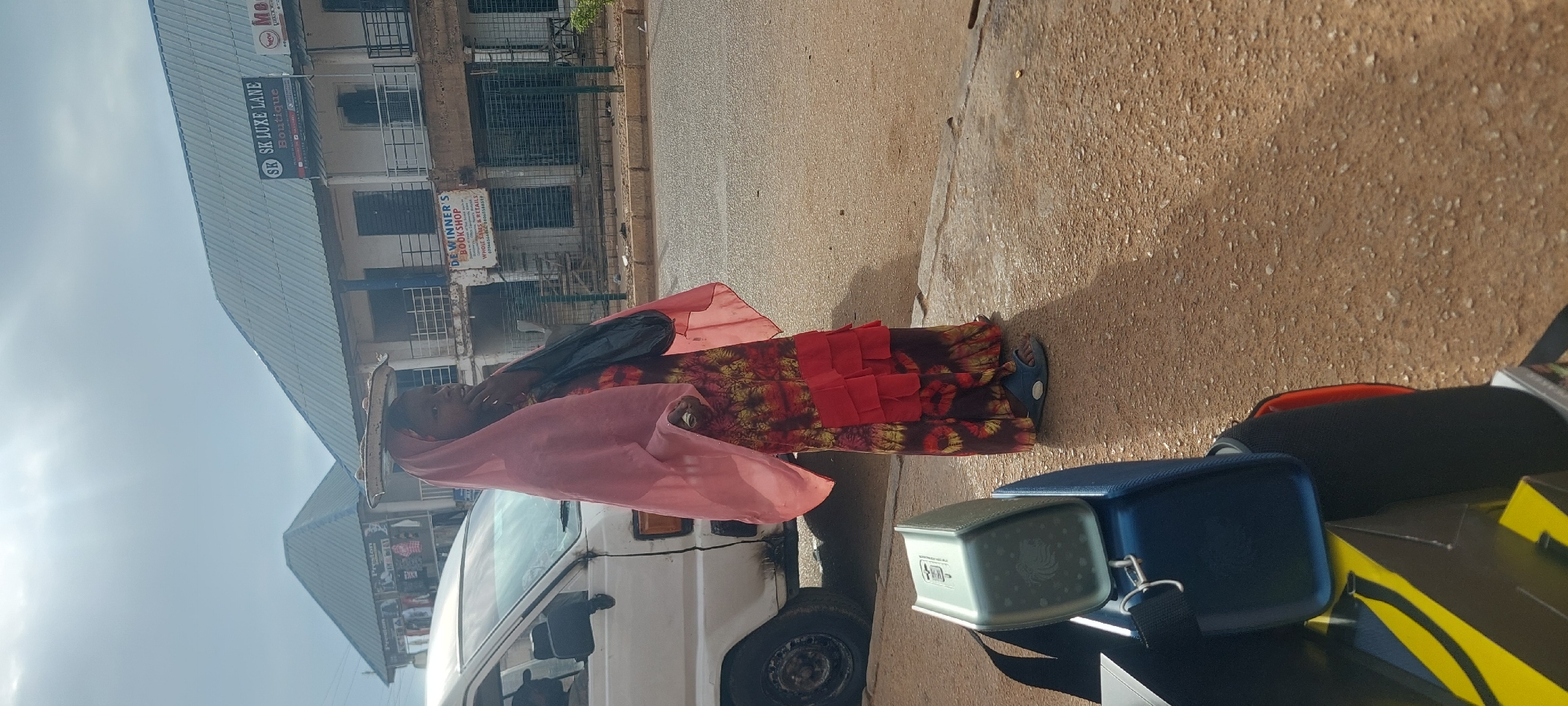 This image captures a daylight street scene in an unknown urban or semi-urban location. **People:** A young woman or girl stands on a paved shoulder or sidewalk on the right side of the frame. She wears a long dress with a dark brown, red, and yellow patterned upper section and multiple tiers of solid red fabric at the hem. A sheer, light pink headscarf (hijab-style) covers her head and drapes over her shoulders. On her feet are blue open-toe sandals with a white detail. She has a flat, round, light-colored tray balanced on her head. Her right hand is raised near her mouth or chin, and her left arm hangs down, holding a small, light-colored object, possibly folded currency. A black shoulder bag is visible across her left shoulder. Her gaze is directed towards the left. **Vehicles:** A white utility van, possibly a Toyota Hiace, is parked or moving slowly on the street to the left of the woman. Only its front left side, including the tire and driver's door, is visible. A person's head is partially discernible inside the driver's side. The van appears worn. **Buildings and Infrastructure:** The scene features a paved road (asphalt/tarmac) and an adjacent paved shoulder/sidewalk separated by a curb. Across the street, there are several multi-story commercial buildings under a mostly overcast sky, with some visible sun rays. - The building directly opposite the woman has a dark blue corrugated metal roof. A prominent sign on its upper level reads "SK LUXE LANE Boutique". Below, on the first floor, another sign reads "DE WINNER'S BOOKSHOP" with smaller text indicating "WHOLE SALES & RETAILS" and two phone numbers (3736654668 and 08035655518). The ground floor features metal security grates or barriers over window/door openings. - To the left of the "SK LUXE LANE" building, another structure with a similar dark blue roof displays signs that include the word "Preston," suggesting a clothing store, with apparel visible through windows. - To the far right, a partial circular sign with "Mo..." or "New" is visible on another building. - Metal railing balconies are present on the upper floors of the buildings across the street. **Foreground Objects:** In the bottom left corner, several objects are partially visible: - A light greenish-grey rectangular portable speaker or electronic device, bearing a lion logo, rests on a dark blue bag or container. - A black strap is attached to the blue object.