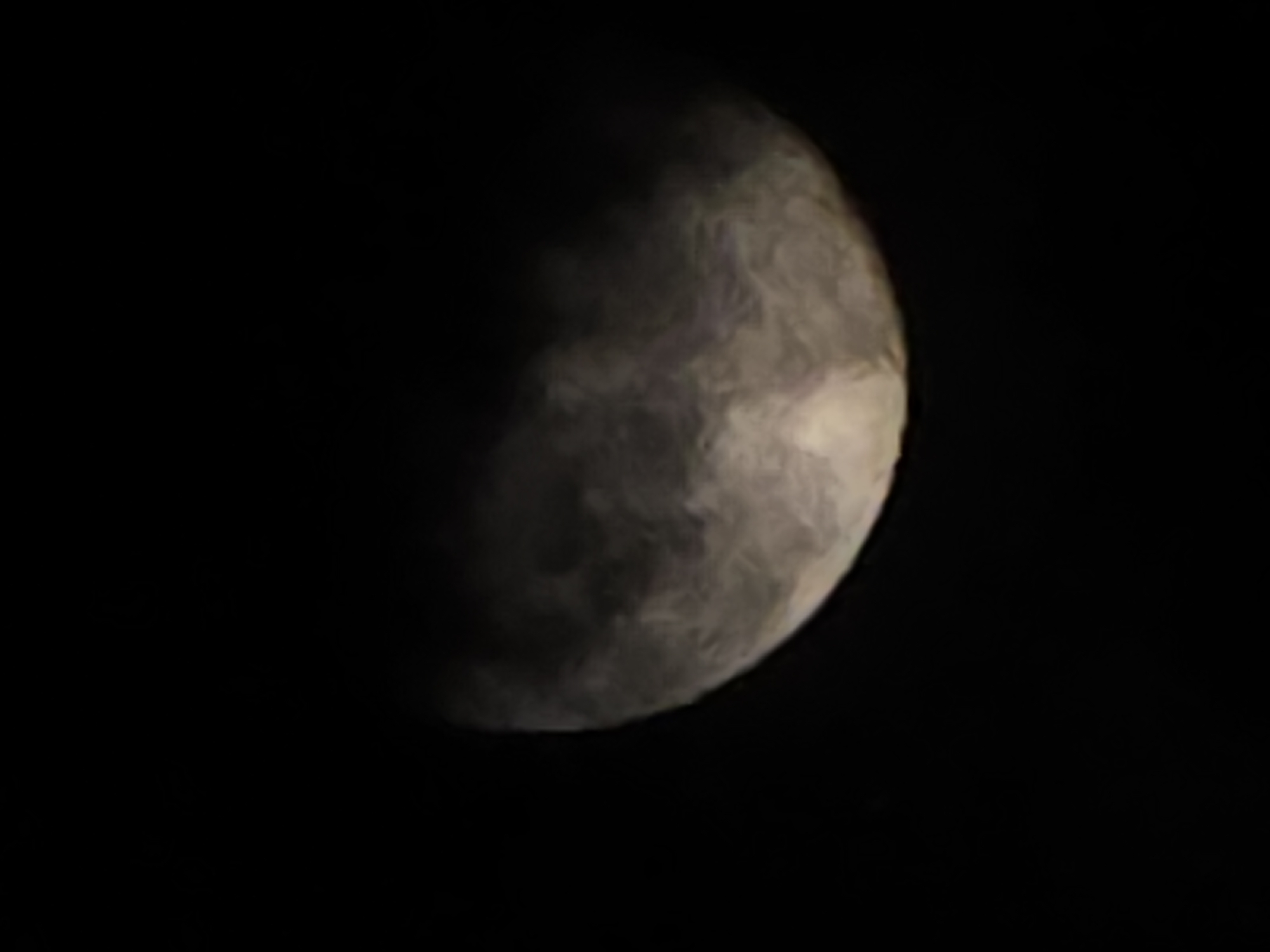 The image is a close-up shot of the moon against a black sky, appearing to be a waning gibbous phase. The moon's surface is visible, with some cloud cover obscuring parts of it, creating a hazy effect. The lighting suggests it is nighttime. There is no indication of the city of Ермолаево, Russia, in the image.