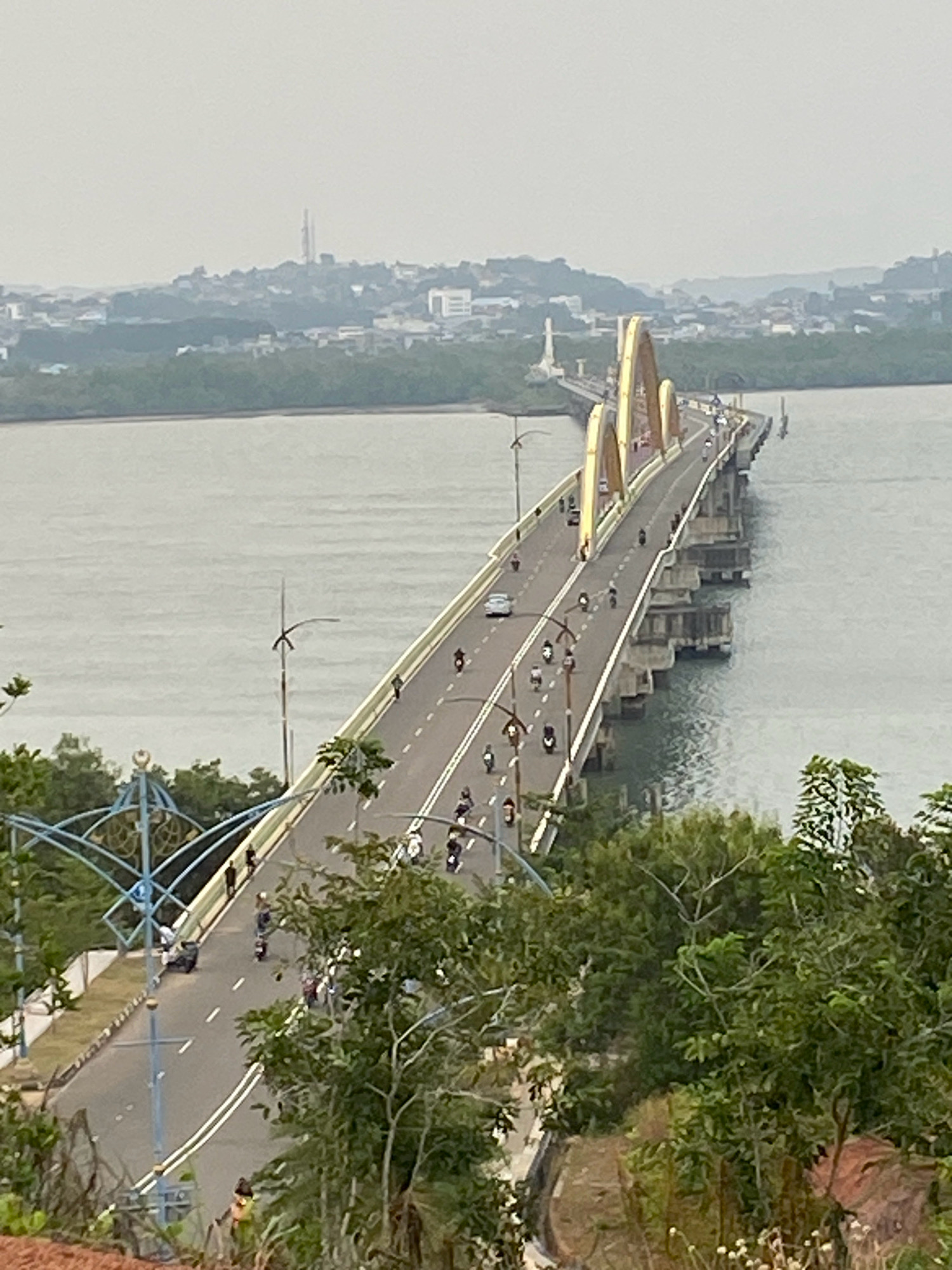 This image captures a view of the Dompak Bridge in Tanjung Pinang, Indonesia, which connects the mainland to Dompak Island. The bridge is a prominent structure, characterized by its distinctive golden arched supports that stand out against the hazy sky. The bridge carries vehicular traffic, including cars and a considerable number of motorcycles, indicating daily use by locals.
The environment surrounding the bridge features a body of water, likely the Riau Strait, with a distant cityscape visible on the horizon. The cityscape appears somewhat obscured by mist or haze, typical of coastal or tropical environments. Lush green vegetation, including trees and bushes, frames the foreground and sides of the bridge, suggesting the presence of natural landscapes near the urban area. The road on the bridge is clearly marked with lanes and white lines. Several streetlights are visible along the bridge's length, and decorative blue metal structures, possibly part of a pedestrian walkway or aesthetic elements, are present on the left side of the bridge's approach. The overall scene depicts a functional piece of infrastructure integrated into a natural and urban setting.