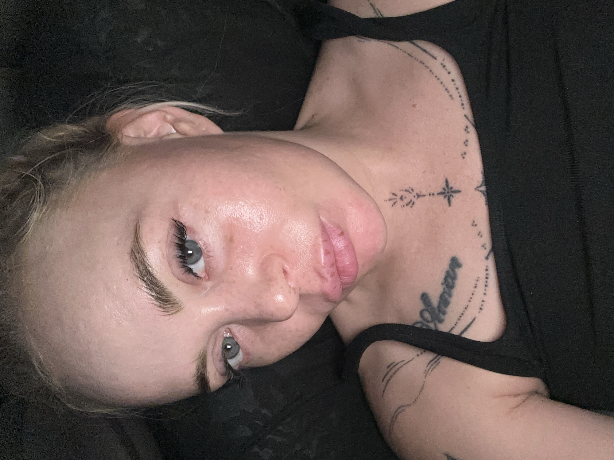A close-up, overhead selfie shows a young white woman with blonde hair pulled back. She has light blue eyes and is wearing false eyelashes. Her forehead and cheeks appear shiny. She has freckles across her nose and cheeks, and her lips are full and slightly parted. She is wearing a black tank top, and a portion of her shoulder and chest are visible. There are several tattoos on her body, including script on her chest and abstract designs on her arm and collarbone. The background is dark and out of focus. The image was taken in an indoor environment, likely a home.