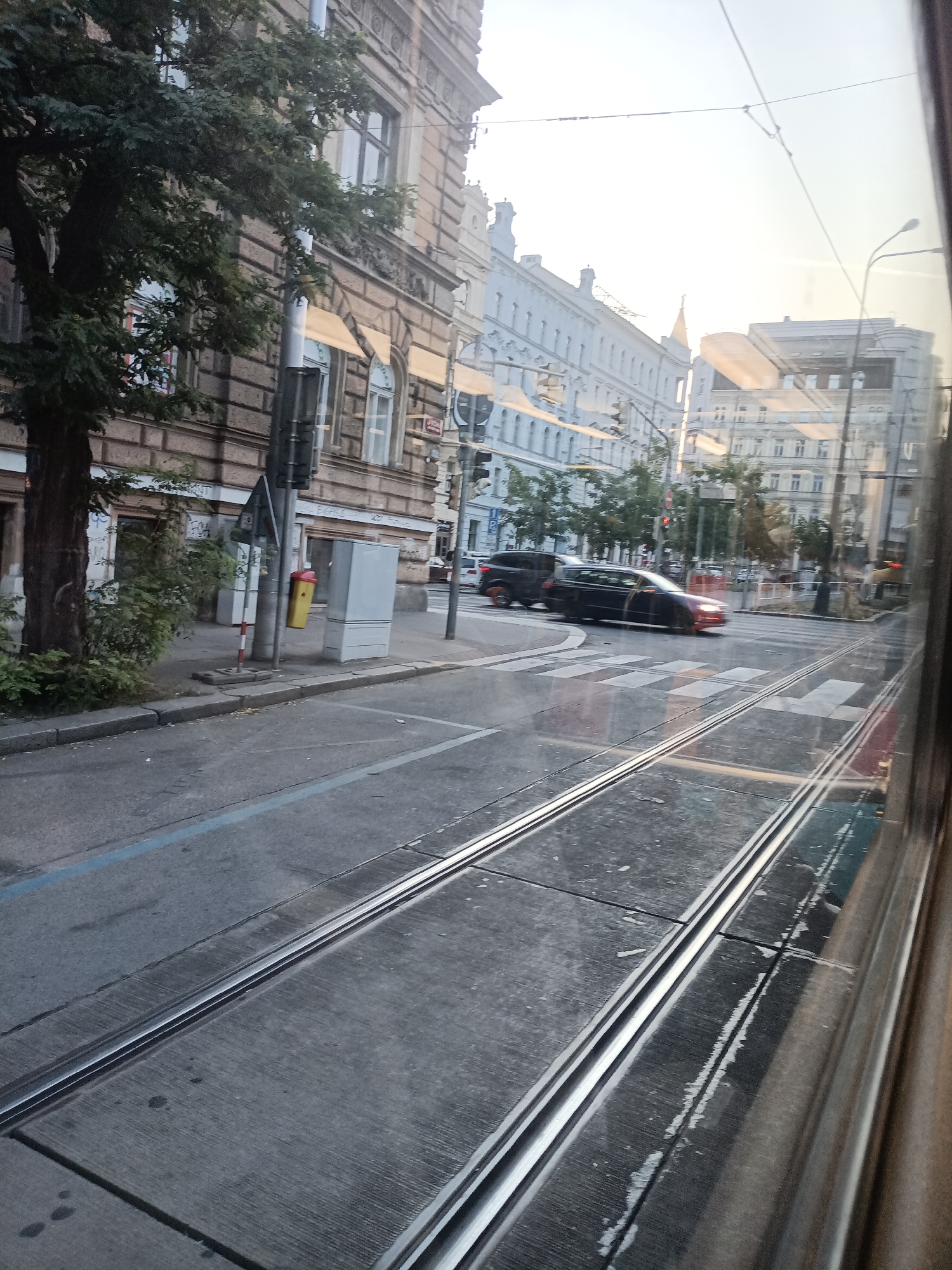The image captures a street scene with tram tracks in the foreground, indicating an urban environment likely within Prague, Czechia, given the architectural style and context. Two dark-colored cars are visible in the middle ground, one of which is a wagon-style vehicle in motion, turning at an intersection marked with a pedestrian crossing. The buildings lining the street feature classic European architecture with ornate facades and numerous windows. A large tree with dense foliage stands on the left side of the street, near a building with a brown, textured facade. Several lampposts and traffic signals are present, along with overhead wires, typical of a city with a tram system. The sky is bright and appears somewhat hazy, suggesting daylight. The perspective is from inside a moving vehicle, likely a tram, looking out a window.
