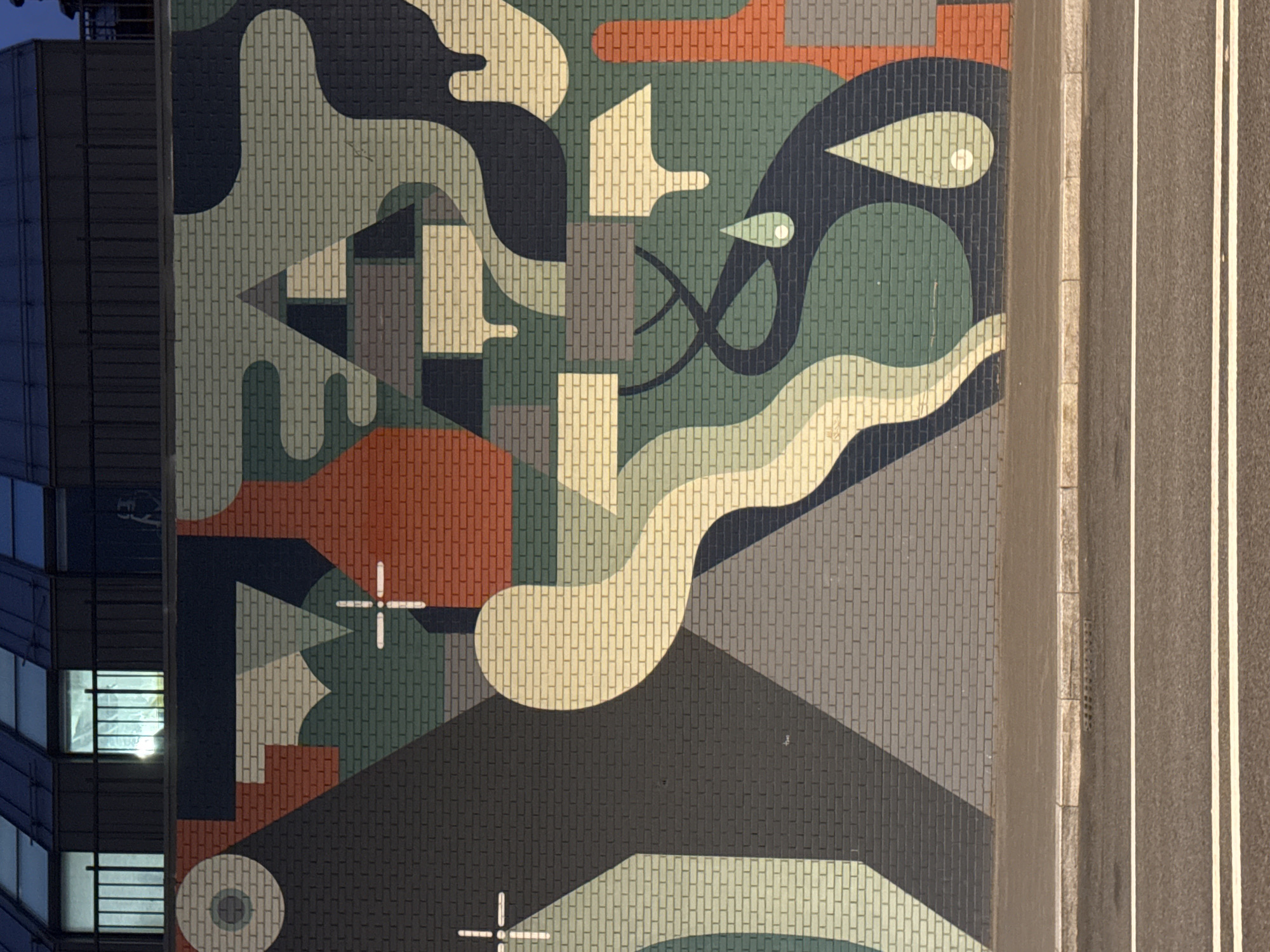 The image is a high-angle shot depicting a mural on the side of a building, adjacent to a road. The mural features an abstract design composed of geometric shapes and curvilinear forms in a palette of muted colors, including greens, blues, grays, and a reddish-brown. The building itself is a modern high-rise with visible window grids. The adjacent road appears to be paved and features double white lines, indicating lanes of traffic. The city or region is unknown.