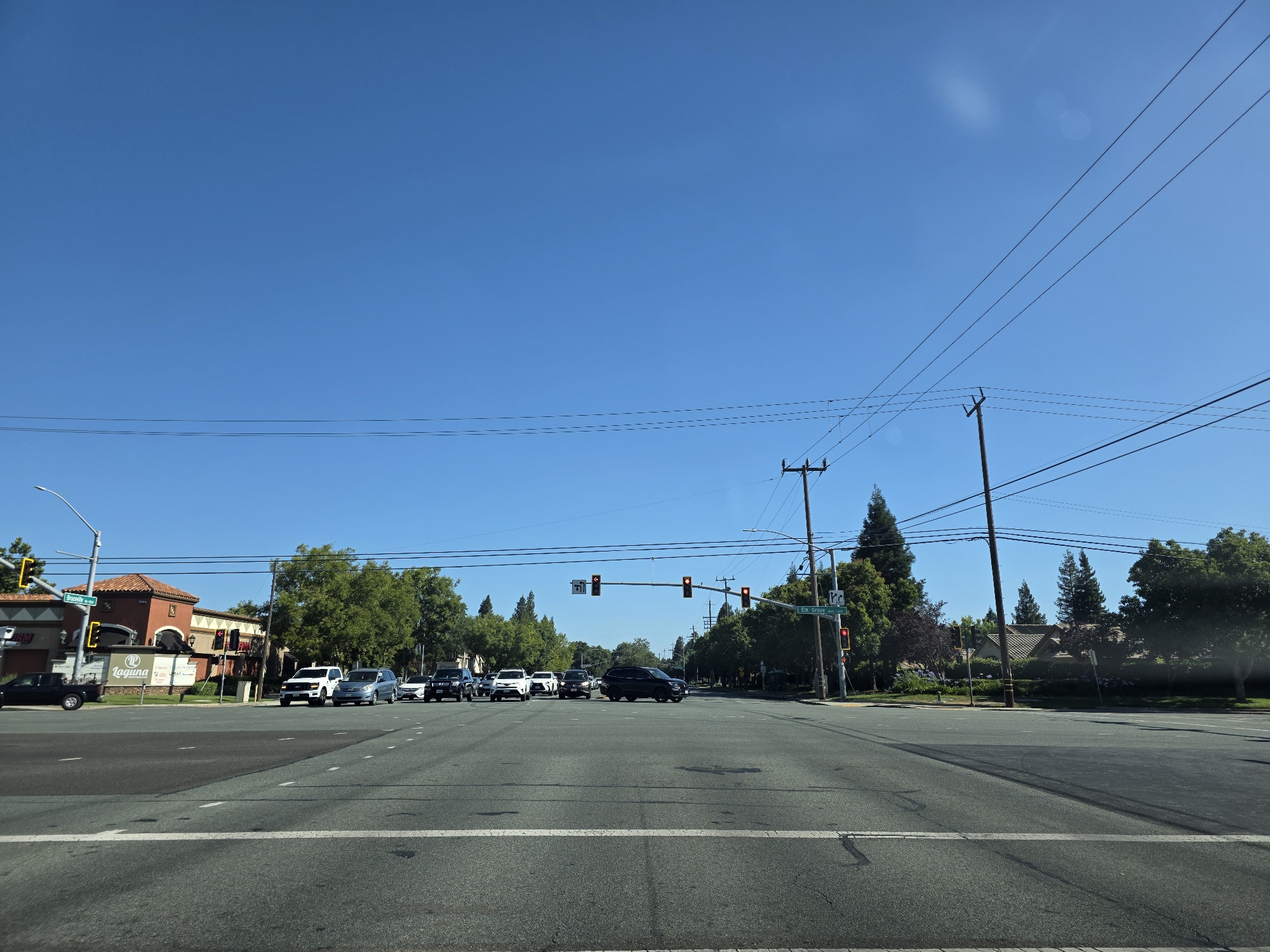 A street scene shows an intersection on a clear, sunny day. Several cars are stopped at a red light, waiting to proceed. A building with a red tile roof and arched windows is visible on the left side of the intersection, with a sign that reads "Laguna" above a logo. Other buildings and trees line the sides of the road. Power lines crisscross the sky, supported by wooden poles. A street sign indicates "Elk Grove Blvd" and an arrow pointing straight, suggesting the location is Elk Grove, California. The time of day appears to be late morning or early afternoon due to the bright sunlight and clear blue sky. No people are visible in the scene.