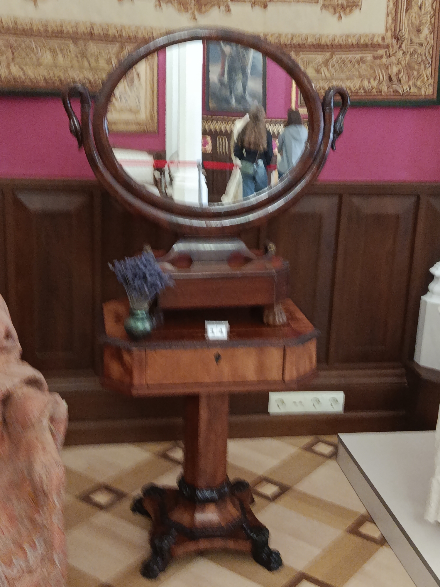 The image depicts an interior scene, likely within a museum or historical building in Kosava, Belarus. In the foreground, a wooden vanity table with a circular mirror stands on a pedestal with ornate legs. The mirror reflects two individuals, one wearing a dark top and blue jeans, and the other in a light gray jacket. A small vase with dried lavender sits on the vanity. In the background, a wall is painted pink and adorned with framed artwork. The floor features a patterned tile design. To the left, part of a sculpture or statue is visible. To the right, a white object, possibly another pedestal or display, is partially in view. The overall impression is that of a preserved historical setting.