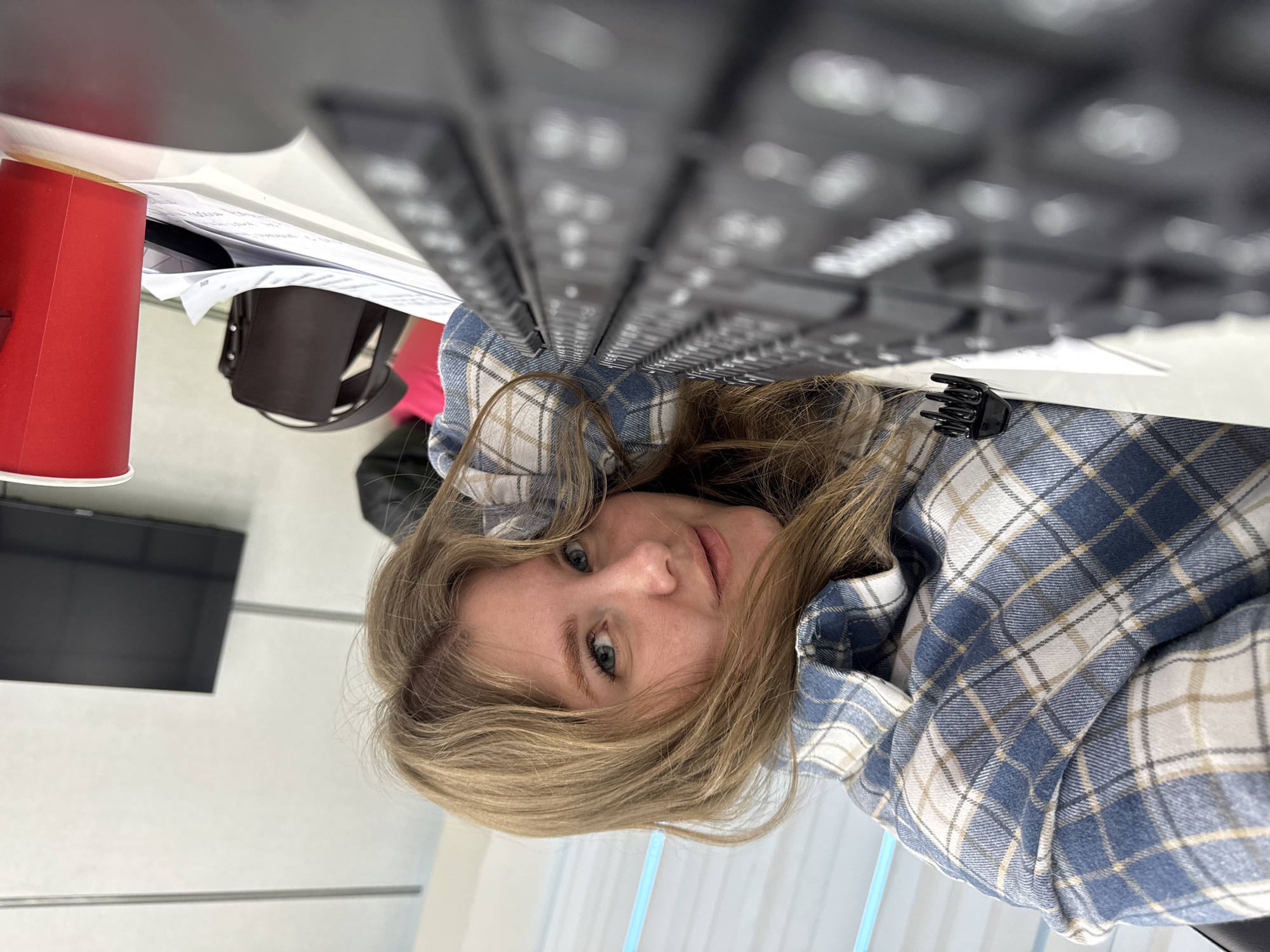 A young woman with blonde hair and blue eyes is lying on a desk next to a black computer keyboard. She is wearing a blue and white plaid shirt. To her left is a red paper cup, papers, and a black purse. In the background is a wall with a black rectangular object, likely a television. The location is likely an indoor office space. There is no indication of the city of Perm, Russia in the image.