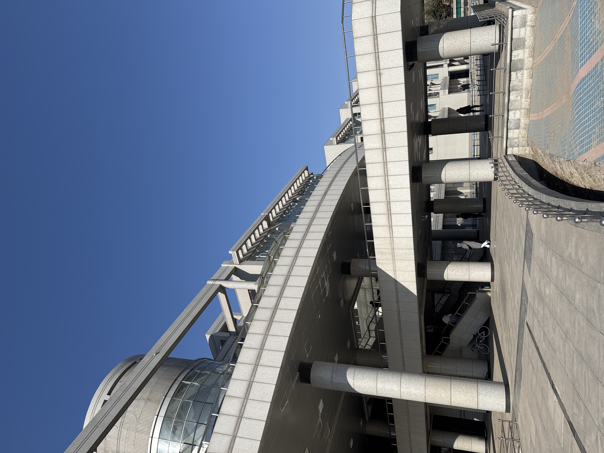 From a low angle, a view of a tall, modern building in Zhongtun, China is presented against a clear blue sky. The building’s exterior features a combination of curved glass facades and segmented, geometric structures constructed from light-colored stone or concrete. The building’s design includes cylindrical columns and visible stairwells, adding to its architectural complexity. Near the base of the building, the surrounding area includes a paved walkway and some greenery. A few individuals can be seen walking along the pathway.