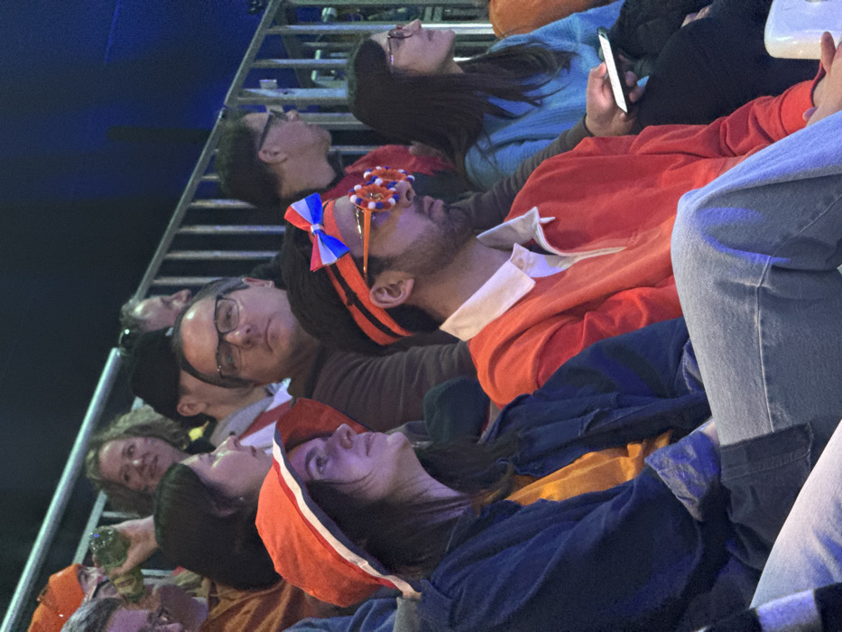 The image depicts a section of tiered seating within an indoor venue in Rho, Italy, populated by multiple spectators. In the mid-foreground, a young woman with dark hair wears an orange bucket hat with white and blue trim, a yellow shirt, and a dark blue denim jacket; her head is turned right, looking slightly upwards. Adjacent to her, a young man with a beard wears an orange long-sleeved polo shirt, an orange headband adorned with a red, white, and blue bow, and orange novelty glasses with multi-colored circular elements. He faces forward. To his right, a woman with dark hair and glasses, clad in a light blue textured sweater, holds an illuminated white smartphone in her right hand and looks to her right. Behind these individuals, a man with glasses in a dark-colored sweater looks directly forward. Several other spectators are visible, partially obscured, throughout the frame, including one woman in the upper left holding a green bottle and multiple individuals wearing orange clothing. A silver metal railing system demarcates seating levels in the background, where the environment is predominantly dark with subtle blue ambient lighting.