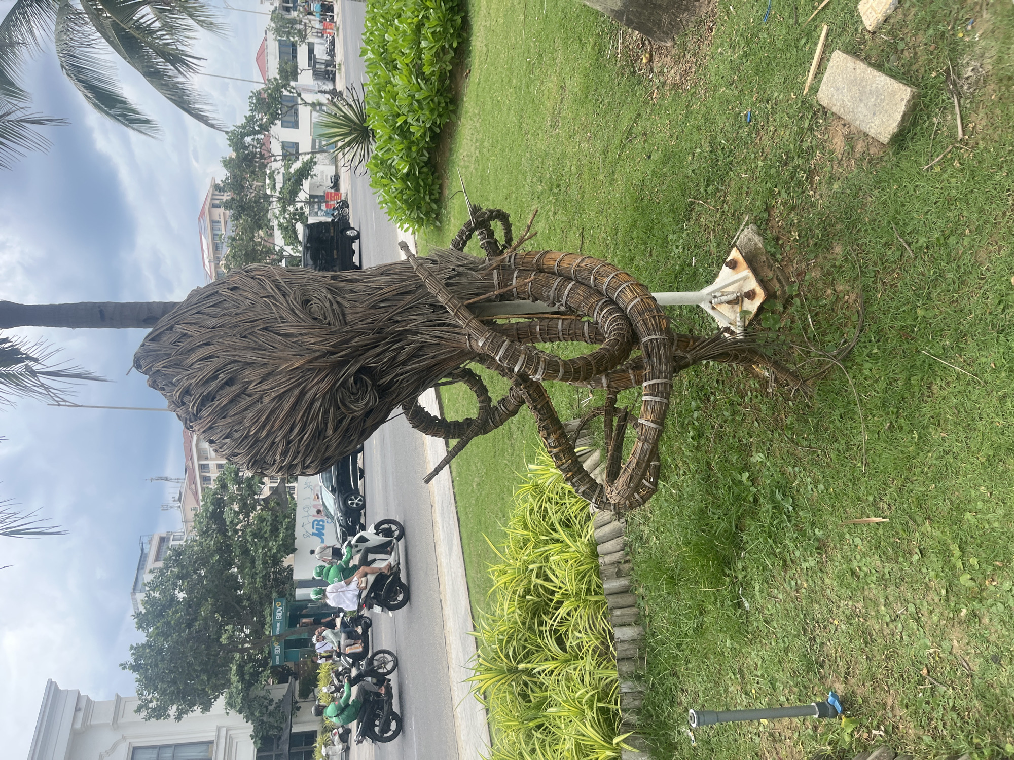 In Đà Nẵng, Vietnam, an outdoor scene features a unique sculpture made of dried palm fronds and bound with wire, resembling an octopus or a similar sea creature. The sculpture stands on a grassy area with a concrete base and metal supports. In the background, a street bustles with activity: several people ride motorcycles, some wearing green uniforms, suggesting a delivery service. A black car is also visible on the road, along with buildings and palm trees lining the street. The sky is overcast. Lush green bushes and a row of decorative plants with yellow and green leaves are planted in front of the sculpture, adding to the urban landscaping.