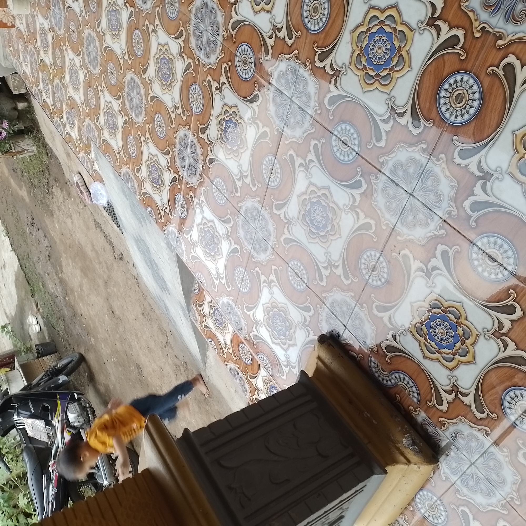 In Jatisobo, Indonesia, a young child in an orange shirt and blue pants is running. The child's body is in motion, captured mid-stride as they move from left to right. A motorcycle is parked to the left of the scene, and a portion of a decorative pillar is visible in the foreground. The ground is covered in patterned tiles, predominantly brown and white with floral motifs. The overall setting appears to be an outdoor area, possibly a patio or walkway, adjacent to a dirt path where the motorcycle is situated.