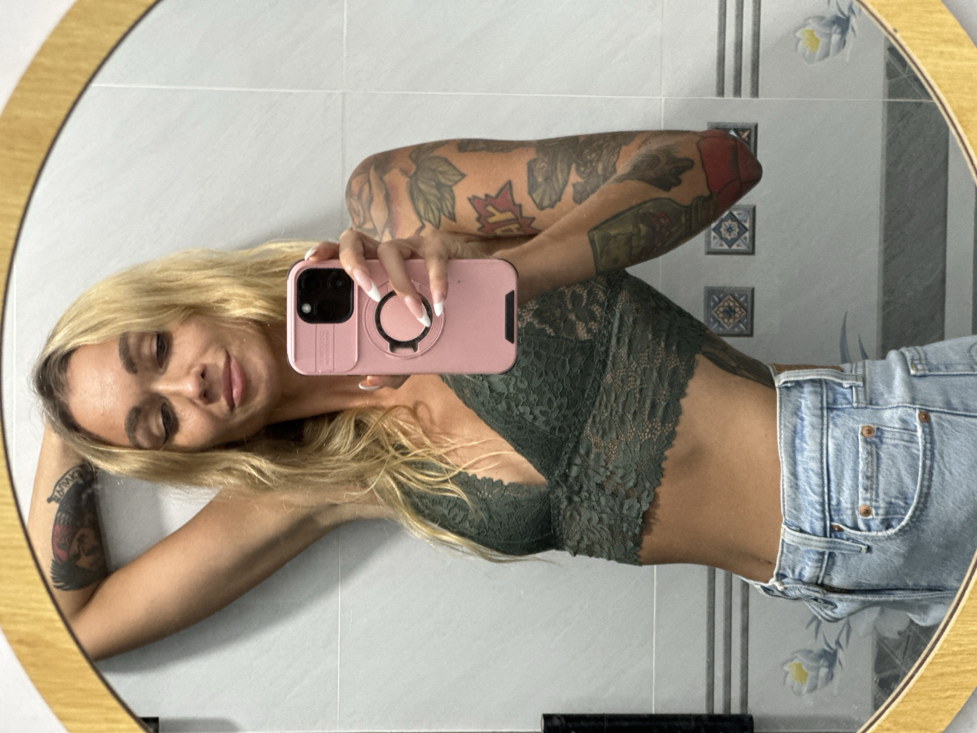 A woman with blonde hair takes a mirror selfie in a bathroom. She is wearing a green lace bralette and light-wash denim jeans. She has multiple tattoos on her arms. The bathroom has white tiled walls with some decorative tiles and a mirror with a golden frame. The scene is likely indoors.
