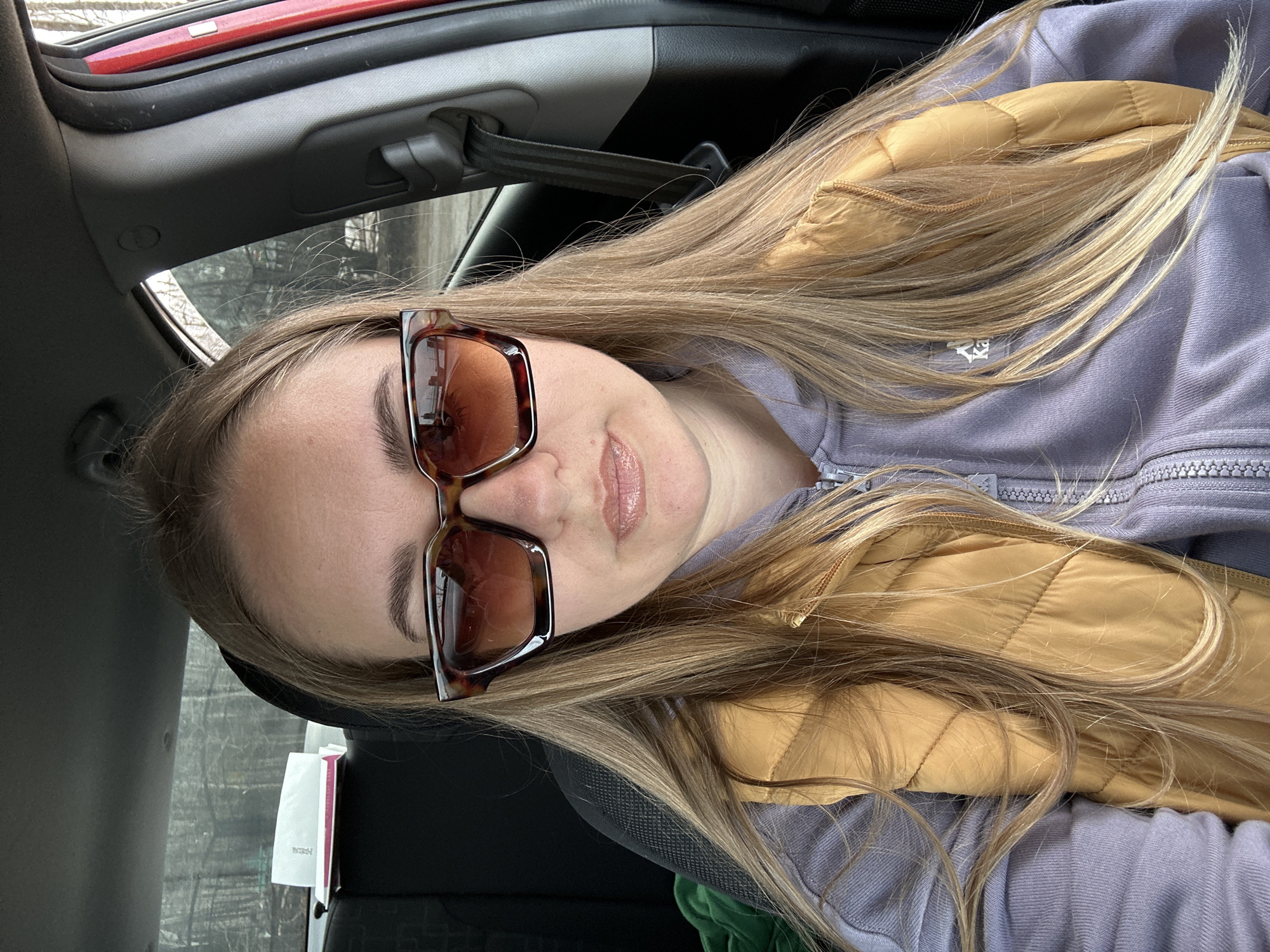 A woman is seated in a car, wearing sunglasses, a lavender-colored zippered sweatshirt, and a golden-brown quilted vest. The interior of the car is visible, including the dashboard and seatbelt. Outside the car window, there are bare trees. Without additional context, it is not possible to confirm the location is Chaikovsky, Russia.